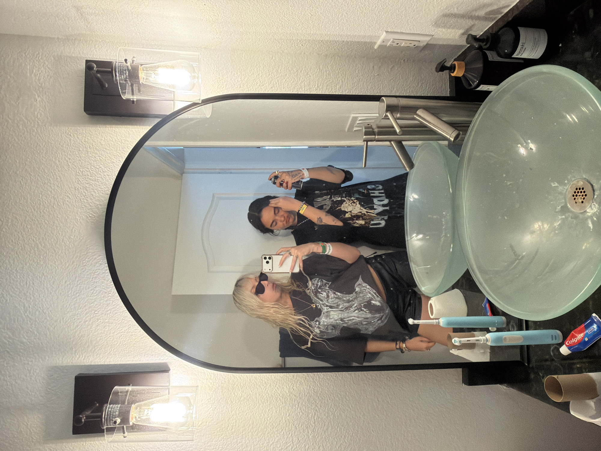 This image shows a reflection in a bathroom mirror. Two people are visible in the reflection, both holding phones and taking pictures of themselves. The person in the foreground has blonde hair and is wearing sunglasses and a dark t-shirt. The person in the background has dark hair and is wearing a dark t-shirt. The bathroom features two vessel sinks, two electric toothbrushes, a tube of toothpaste, and two black pump bottles. The location is Palm Desert, United States of America.