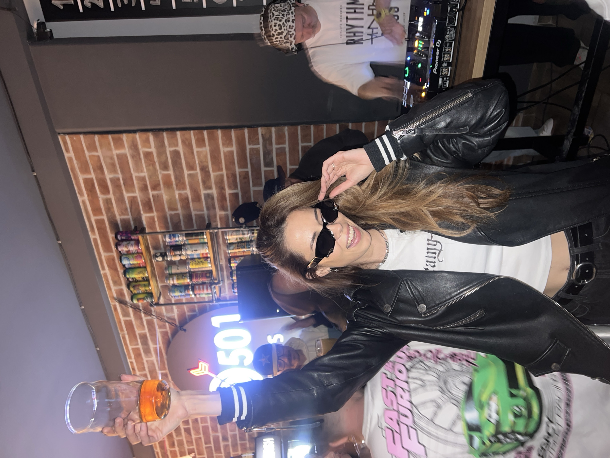 An indoor scene is depicted within what is identified as 501 Pub Bar in Bar, Montenegro. The central figure is a woman with long, light brown hair, positioned slightly to the left of the frame's center. She wears dark, wave-framed sunglasses, a white crop top with partially visible text, a silver chain necklace, a black leather motorcycle jacket with striped cuffs on her left arm, and dark pants with a black belt and silver buckle. Her left arm is raised, holding a clear glass containing an amber liquid, approximately one-quarter full. Her right hand is positioned near her right temple, appearing to adjust her sunglasses, and she displays a wide smile.
Behind her, a textured brick wall extends across the background, partially obscured by other individuals and objects. To the left of the woman, a blurred individual wears a white t-shirt with "FAST FURIOUS" text and a graphic of a green car. Above the woman's left shoulder, a circular neon sign displays the number "501" in white light with a red arrow pointing left. To the right of the neon sign, two industrial-style shelves mounted on the brick wall hold multiple rows of assorted beverage cans.
Further to the right, a male DJ is visible, operating black Pioneer DJ mixing equipment set on a wooden surface. He wears a white t-shirt with "RHYTHM" text and a leopard print bucket hat. Other blurred figures are present in the background, including a man behind the central woman wearing a dark baseball cap with a star logo and glasses, holding a glass. The ceiling is plain and light-colored.
