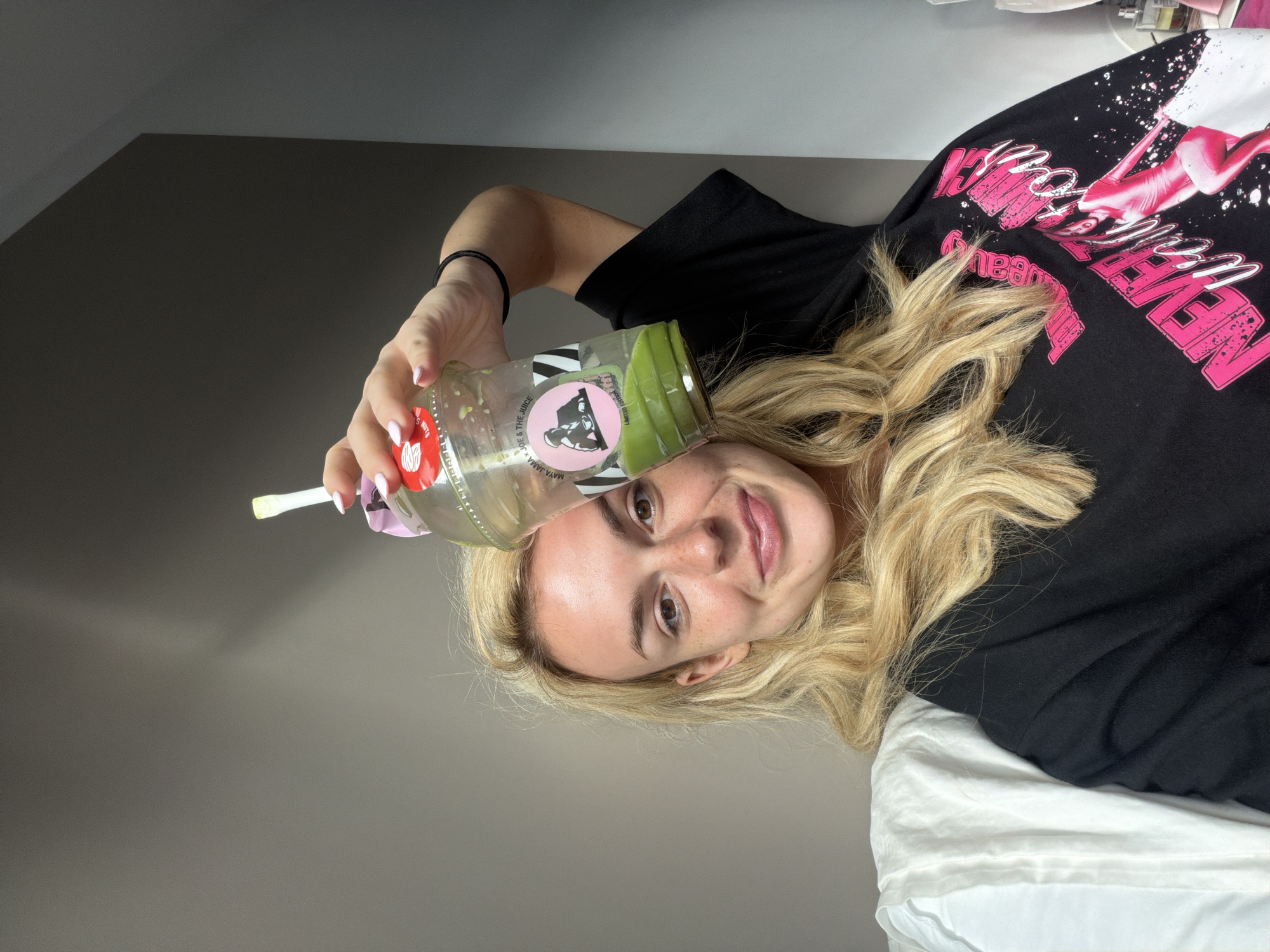 The image features a young woman with blonde hair, light skin, freckles, and brown eyes, lying down and looking directly at the camera with a relaxed smile. She is holding a clear plastic bottle containing a green liquid and a white straw in her left hand. The bottle features a pink and black label with the text "MAYA JAMA x JOE & THE JUICE," "LIMITED EDITION," and "THE SHINE" clearly visible, indicating a branded juice or smoothie.
She is wearing a black t-shirt adorned with vibrant pink and white graphics. Prominent text on her shirt includes the word "NEVER" in large pink letters, along with part of a graphic depicting a figure in a pink outfit. Her blonde hair is spread around her head and rests on white bedding, which is visible on the right side of the frame. The background is a plain, dark grey wall, suggesting an indoor setting, likely a bedroom. The overall mood is casual and relaxed. The location context for the image is London, United Kingdom.