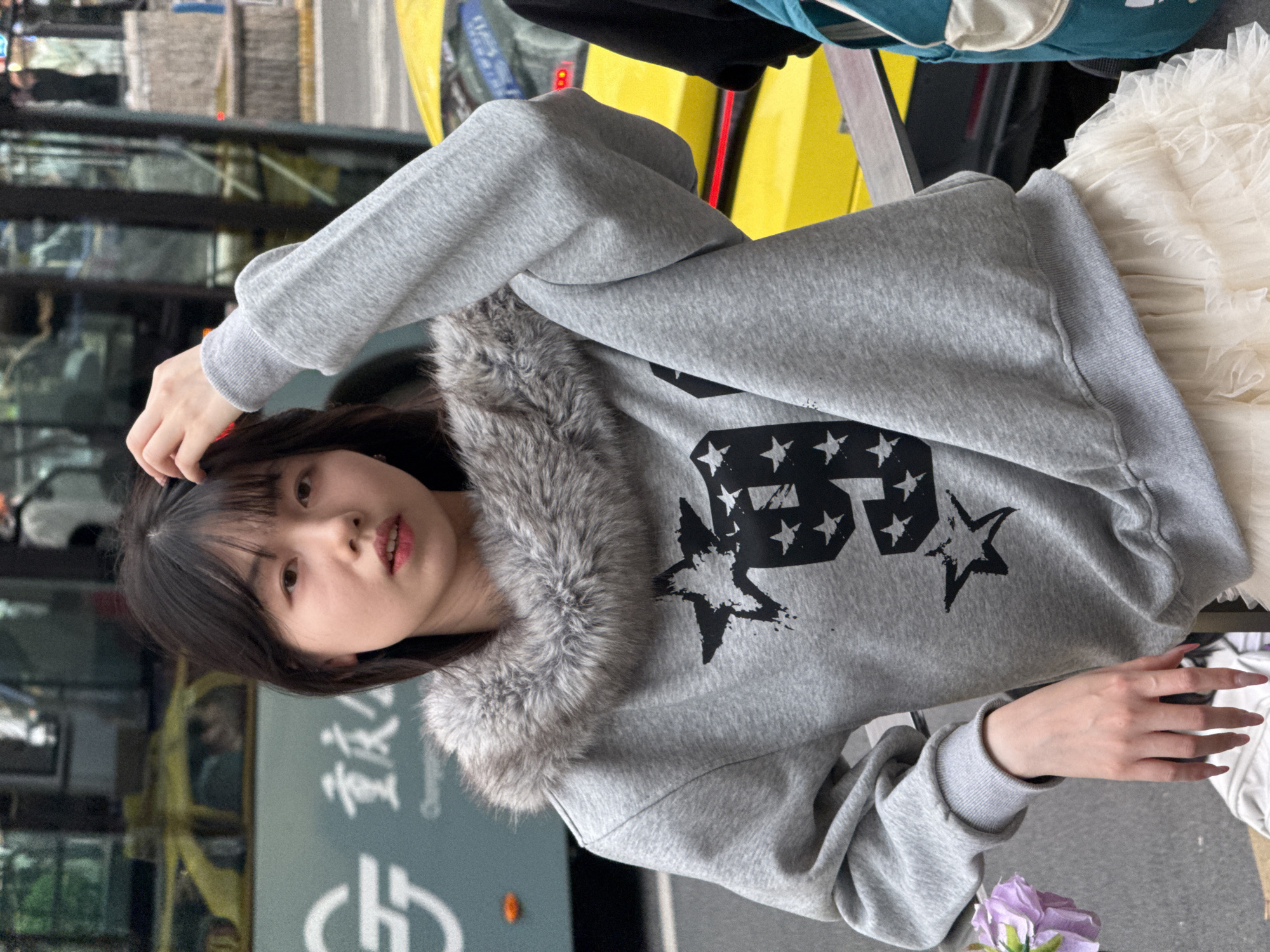 A young woman is seen in an urban setting, standing near a street, possibly in Liangjiang, China. She is wearing a gray sweatshirt with a fur collar and an American flag design, along with a white tulle skirt. The woman has dark hair with bangs and is seen holding flowers. She is standing near a bus and a yellow taxi cab.