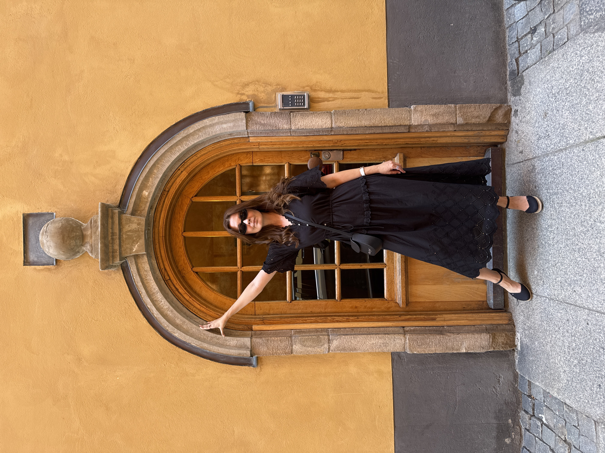 A woman is standing on a stone-paved sidewalk in front of a large, arched wooden door, set into a textured, ochre-colored wall. She is wearing a black midi dress with short, ruffled sleeves and a decorative hem, black espadrilles, and sunglasses. A small black crossbody bag hangs from her shoulder. Her left arm is extended upwards, touching the stone frame of the doorway. To the right of the door, a silver intercom panel is mounted on the wall. The scene appears to be in the Gamla Stan district of Stockholm, Sweden, identifiable by the historic architecture and cobblestone paving.