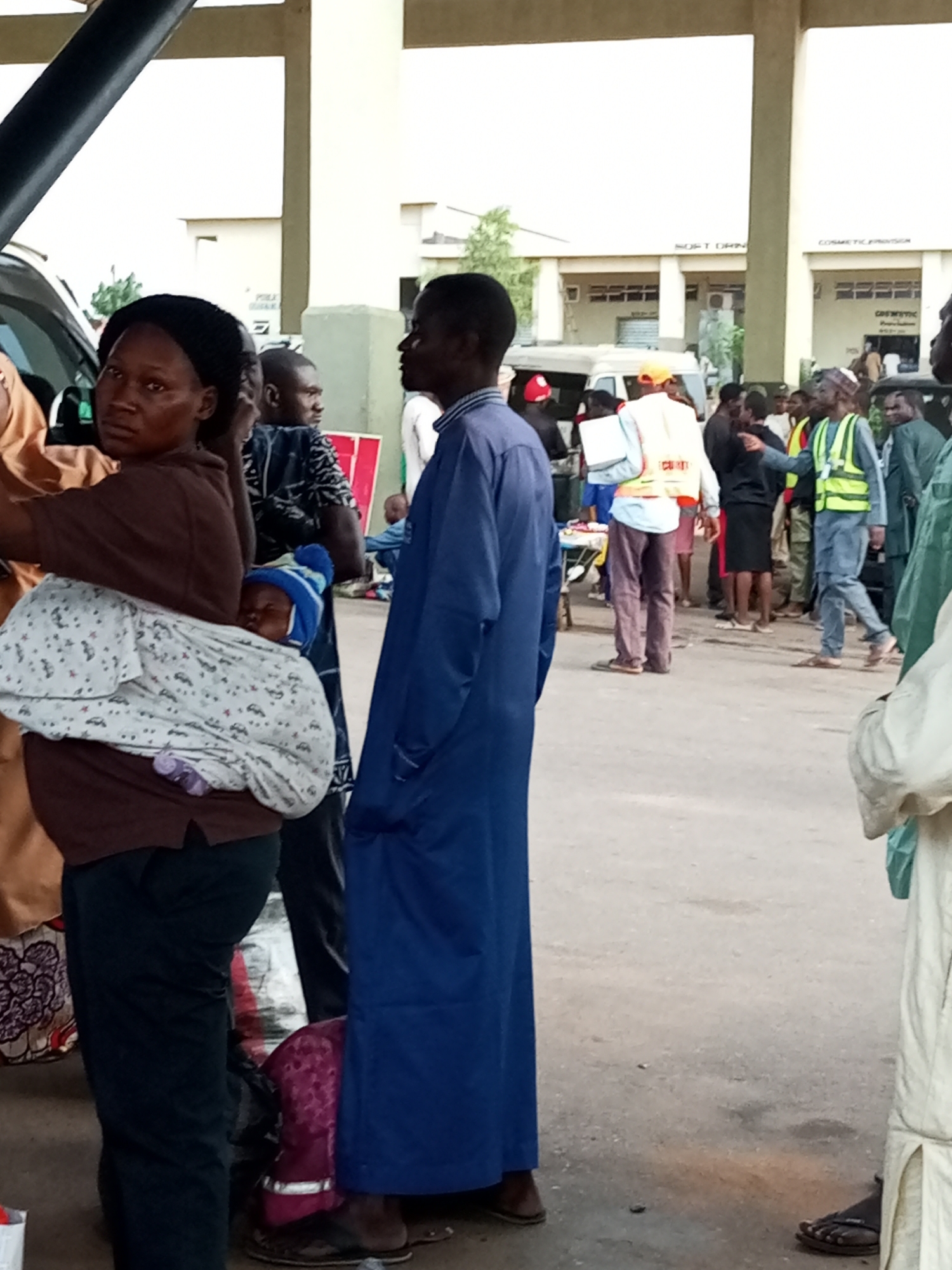 The image depicts a crowd of people in what appears to be a public space in Gombe, Nigeria, likely near a transportation hub given the presence of vehicles. The scene is bustling, with individuals of various ages and genders. In the foreground, a woman is carrying a baby wrapped in a patterned cloth, with the baby's head wearing a blue and white striped hat. She is looking towards the camera. To her right, a man in a blue robe stands, facing away from the viewer. Further into the background, several other individuals are visible. One man wears a bright yellow safety vest over an orange shirt and appears to be a security guard or official, holding a white folder. Another person in a similar yellow vest is also present. The environment includes paved ground and buildings in the background, one of which has signage that is partially legible, suggesting commercial activity such as "SOFT DRIN" and "COSMETIC PROVISION". The overall atmosphere suggests a moment of transit or gathering in a busy urban area.