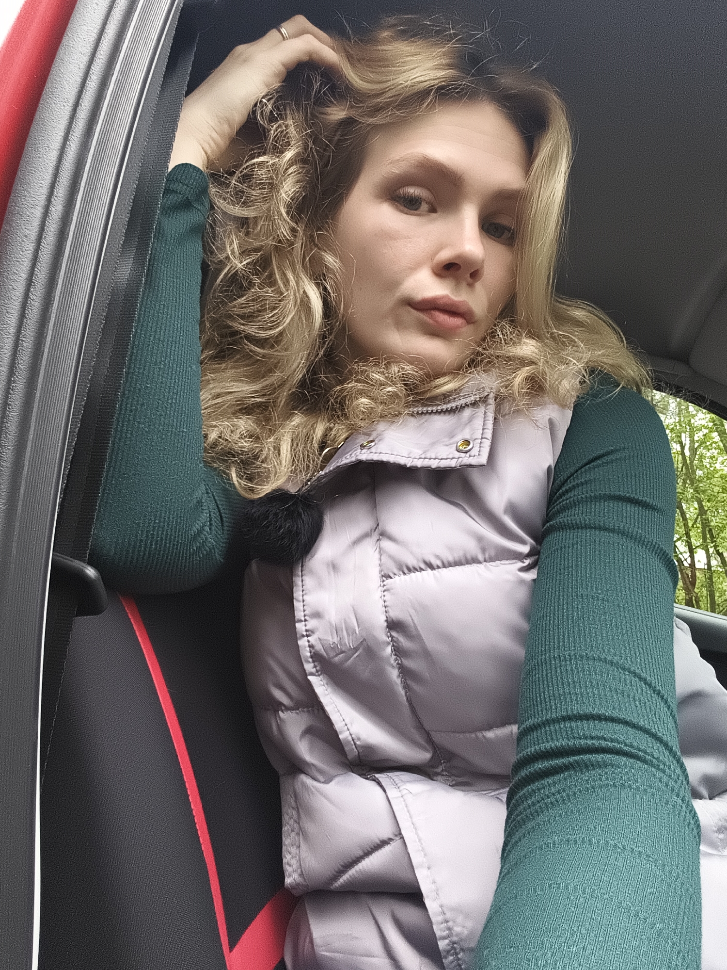 A blonde woman with curly hair sits in the front seat of a car. She wears a silver puffer vest over a green long-sleeved shirt. Her right arm is raised, hand touching her hair. The car interior is visible, including the seat and door. Outside the window, green trees suggest a wooded or park-like setting. The city of Izhevsk, Russia, cannot be determined from the image.