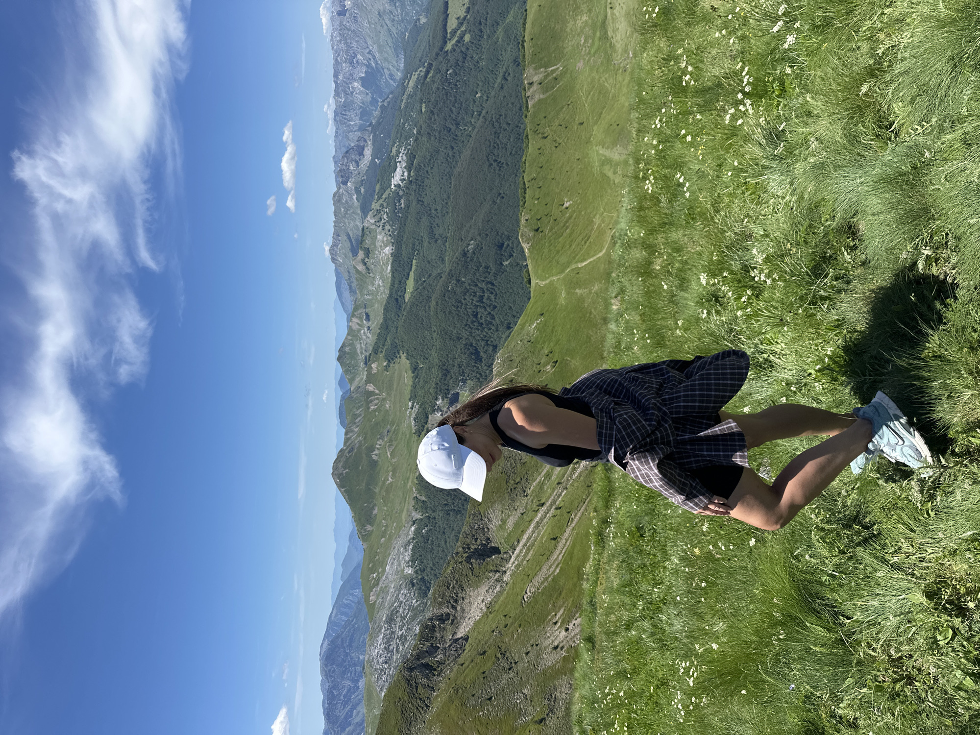 A young woman wearing a white baseball cap, a black tank top, and shorts is standing on a grassy slope with a plaid shirt tied around her waist. She is looking down and to her left. The background features a vast mountainous landscape with green hills and valleys under a bright blue sky with scattered clouds. The scene appears to be in a natural, outdoor setting, likely in a mountainous region. The specific location within Dolja, Montenegro, is not evident from the image itself, but the visual elements suggest a scenic mountain environment.