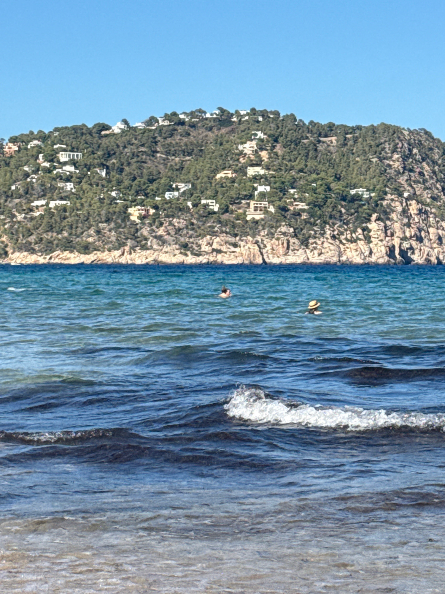 This is an outdoor, medium shot photograph taken during the day. The scene depicts a view of the sea with a hilly, forested coastline in the background. The coastline is dotted with numerous white buildings, suggesting a residential area. The sea is a deep blue with visible waves and a small white crest in the foreground. Two individuals are visible in the water; one person is swimming further out, while another, wearing a straw hat, is closer to the shore. The sky is a clear, bright blue. The location is likely the coast of Santa Eulària des Riu, Spain, given the characteristic Mediterranean landscape with houses built into the hillsides overlooking the sea.