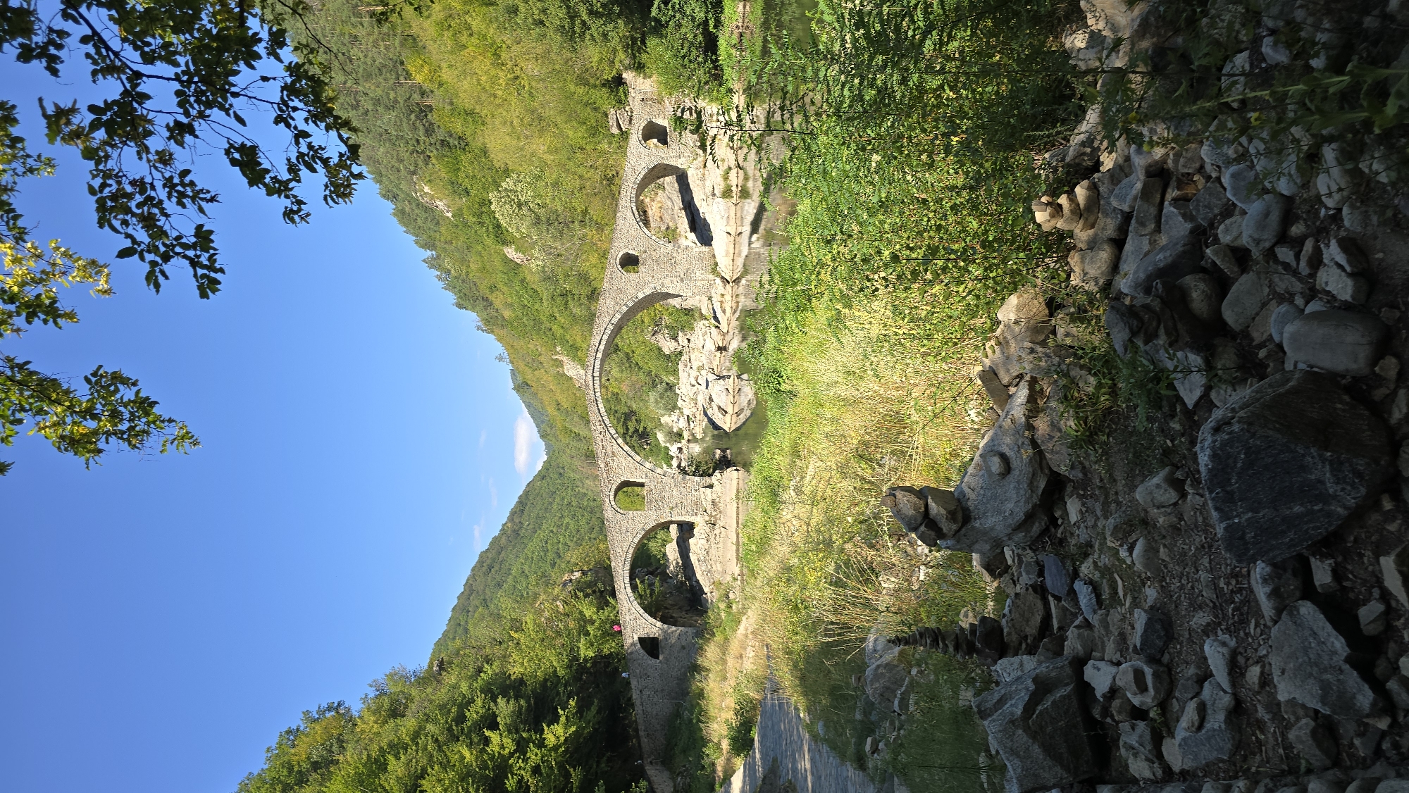 The image shows the "Devil's Bridge" located in the Arda River valley near the village of Dyadovtsi, Bulgaria. The bridge is an ancient stone structure with multiple arches, spanning across a calm river. The surrounding environment is characterized by lush green hillsides covered in trees, and the riverbanks are rocky and overgrown with vegetation. In the foreground, there are several cairns, which are stacks of stones, placed on the rocky ground. The sky is clear and blue. The bridge appears to be a historical landmark and a popular tourist attraction.