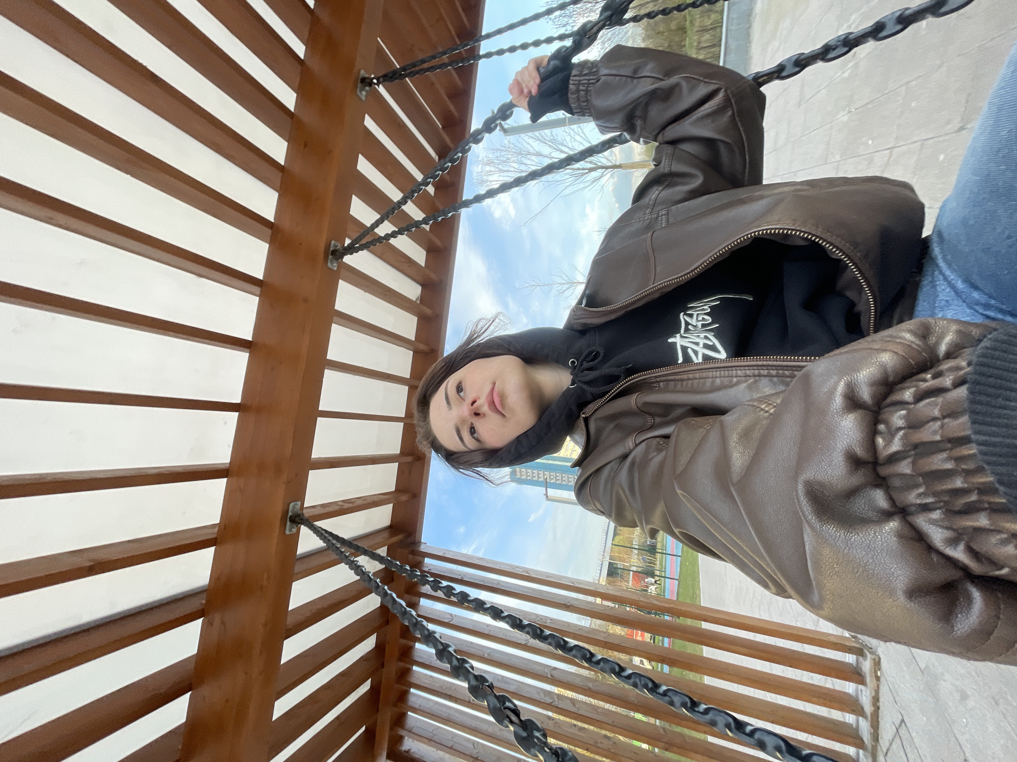 A woman is sitting on a swing in a park in Moscow, Russia. The swing is made of wood and metal chains, and the woman is holding onto the chains with her hands. She is wearing a brown leather jacket, a black hoodie, and jeans. The park is in the background, with trees, grass, and a building visible in the distance.