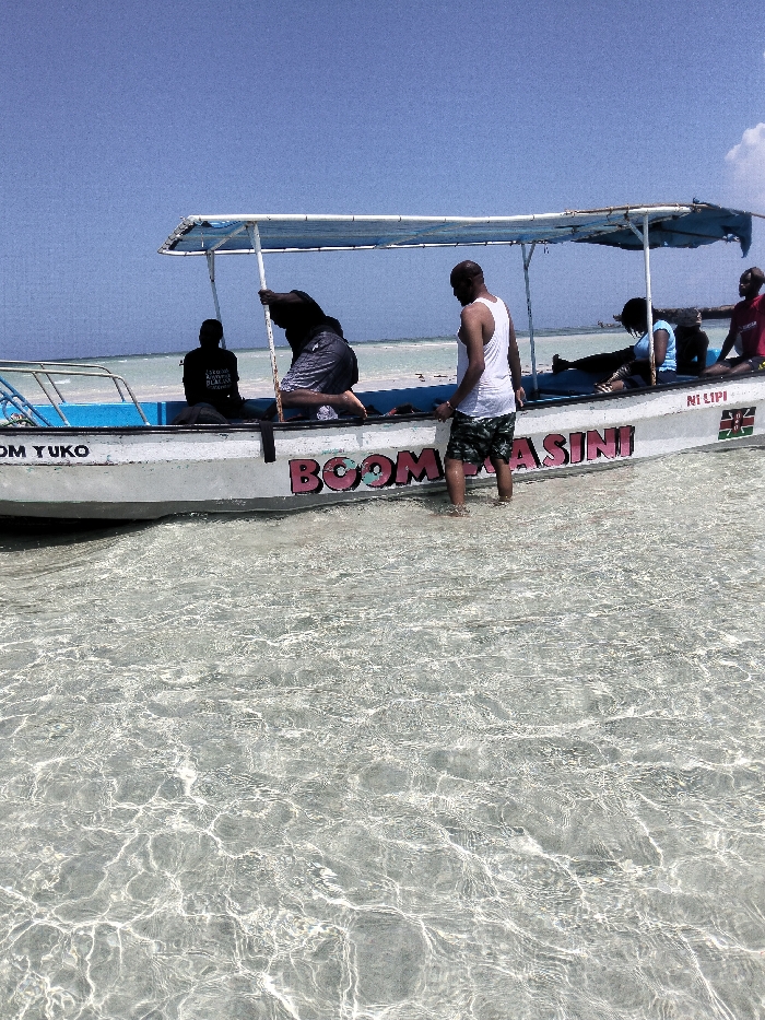 The image depicts a white boat in shallow, clear water under a clear blue sky in Wasini, Kenya. The boat features a blue interior rim and a light blue canvas canopy supported by a metal frame. Pink lettering on the boat's side spells "BOOM WASINI," with "OM YUKO" on the front left and "NI LIPI" alongside a Kenyan flag graphic on the right.
Five individuals are visible. In the immediate foreground, an adult male with dark skin and a bald head, wearing a white tank top and camouflage shorts, stands in the water, facing left towards the boat. His feet are submerged. Inside the boat, to the left, another adult male with dark skin, wearing a dark shirt and patterned shorts, is partially bent over, holding a vertical pole which extends into the water. Further left and seated, another individual in a dark shirt faces away from the camera. Towards the right side of the boat, a person in a blue top and dark trousers is seated, leaning back, while another individual in a red t-shirt is seated closer to the stern. The water is highly transparent, revealing a light-colored, possibly sandy, seabed. The background consists of calm, light blue ocean extending to a distant horizon, with a small cluster of white clouds in the upper right sky.