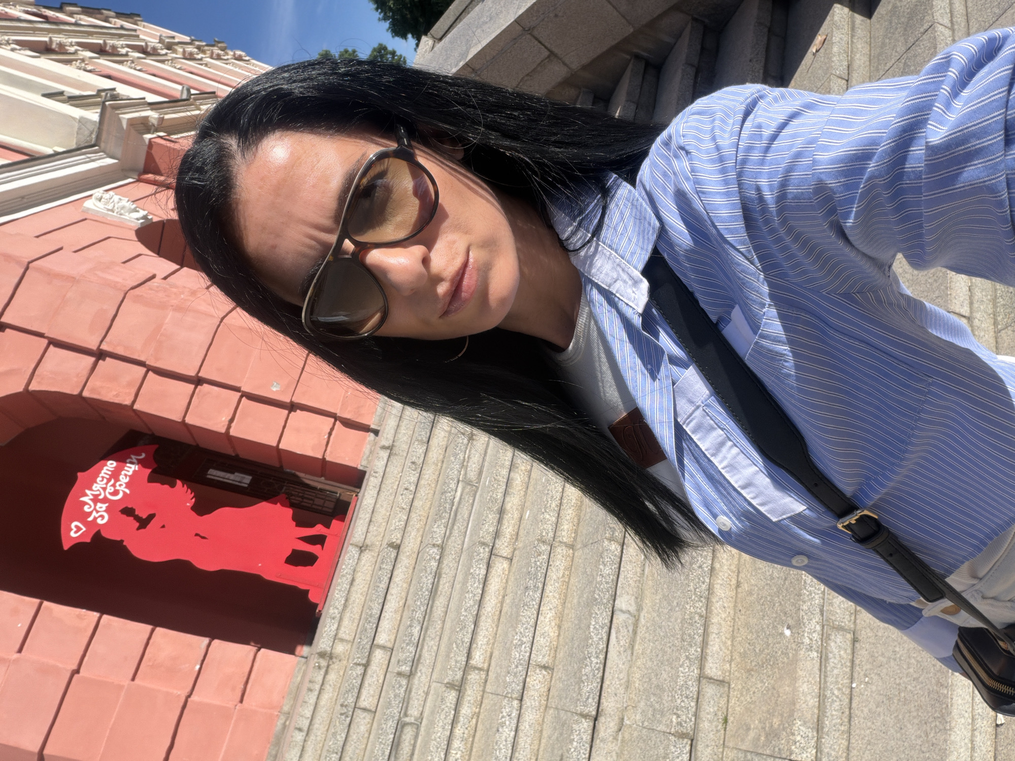 A woman with long, dark hair and sunglasses stands outside a pink building with a red sign in Pleven, Bulgaria. The sign shows a couple embracing under an umbrella with Bulgarian text. The woman is wearing a light blue striped shirt, a white t-shirt, and a black shoulder bag with a gold buckle, and is standing on stone steps. The sky is blue.