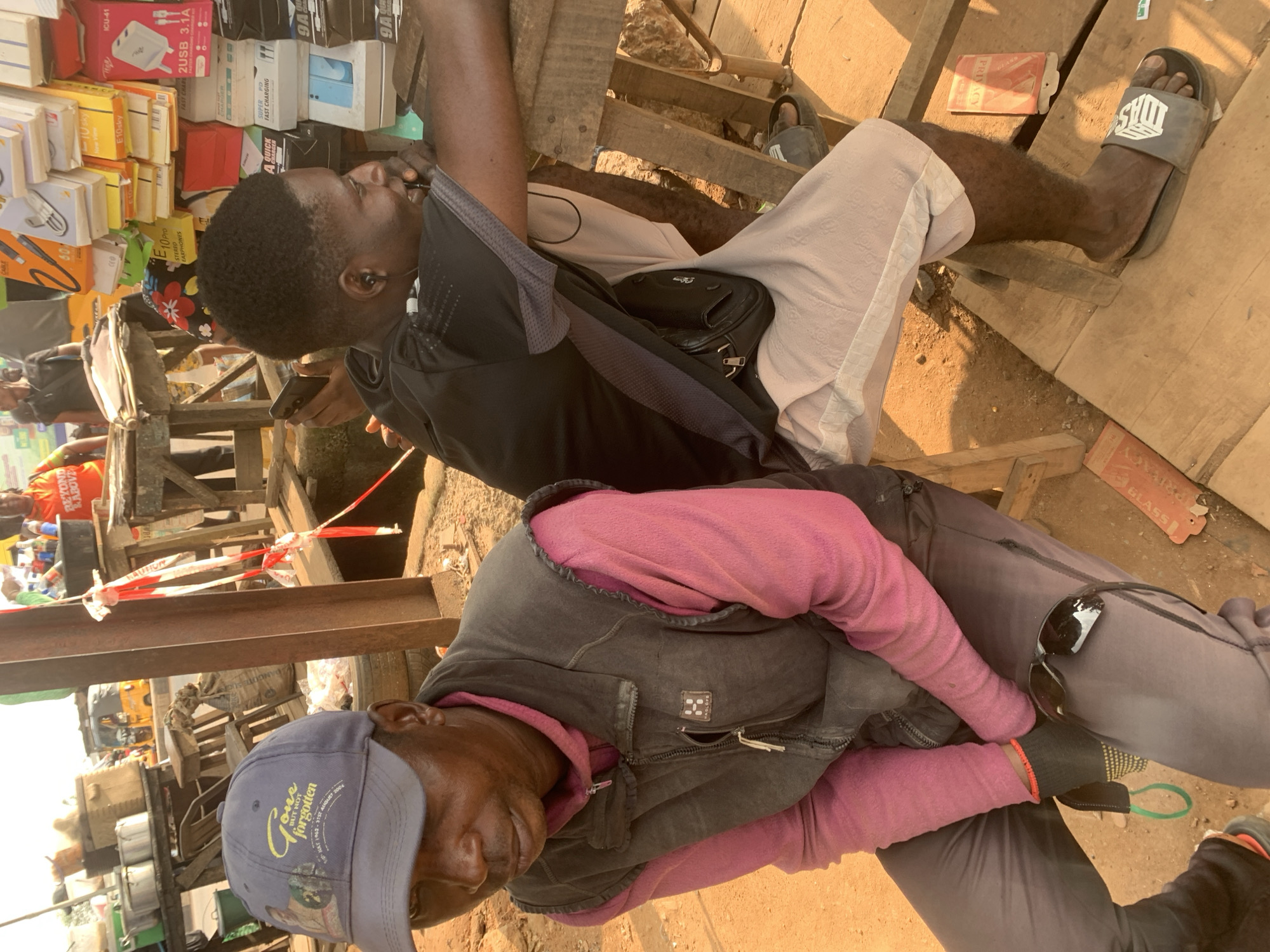 The image depicts a street scene in Shagamu, Nigeria, likely a marketplace. In the foreground, two men are seated on wooden benches. The man on the left wears a grey baseball cap, a pink long-sleeved shirt, and a dark vest. He is looking down and to his left. A pair of sunglasses rests on his right thigh. The man on the right wears a black t-shirt and light-colored shorts. He is looking to his right and appears to be talking on a mobile phone, which he holds in his right hand. He has black earbuds in both ears. A black fanny pack is worn across his waist. The background shows stalls with various items for sale, including what appear to be electronic accessories and household goods. A yellow tricycle is visible in the distance, suggesting local transportation. The ground is a mix of dirt and wooden planks. The overall impression is of a busy outdoor commercial area.