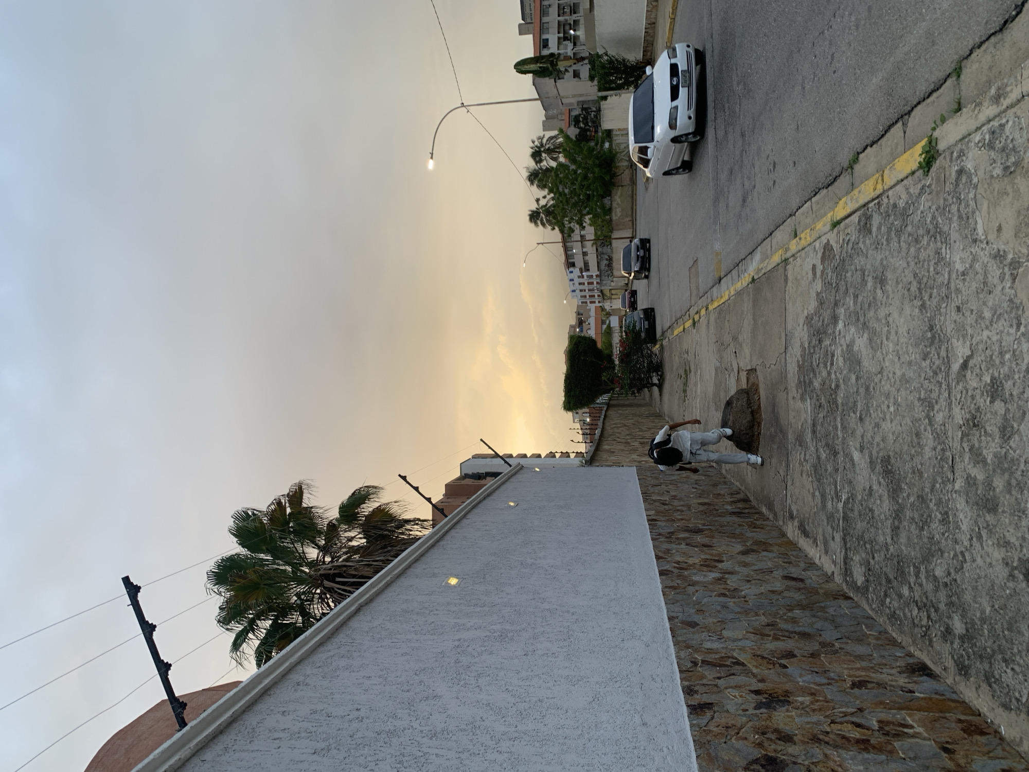 The image depicts an outdoor urban street scene in Pampatar, Venezuela, captured during either sunrise or sunset, indicated by the warm, yellowish-orange glow along the horizon transitioning to a lighter blue-grey sky above. The illuminated streetlamp on the left further suggests the transitional time of day.
In the foreground, a textured white wall with embedded rectangular lights runs diagonally across the lower left. Above this wall, a security fence with multiple wires is visible. A large palm tree with green fronds stands prominently to the left of the wall, silhouetted against the sky.
The main subject of the scene is a paved asphalt street receding into the distance. A bright white sedan, possibly a Nissan, is parked on the right side of the street, facing away from the viewer. Several other cars, darker in color, are visible further down the road. A tall streetlamp with a glowing bulb provides light on the left side of the street.
Adjacent to the street is a concrete sidewalk showing signs of age, with cracks and small patches of green vegetation growing from its edges. A yellow painted curb delineates the sidewalk from the road. A manhole cover is visible on the sidewalk in the mid-ground.
To the right of the sidewalk, a person wearing a white top, light pants, and a dark cap is seen walking on a path made of irregular stone or cobblestones. They are positioned next to a tall, rough-hewn stone wall that forms the boundary of an elevated area.
In the background, numerous multi-story buildings with visible windows and balconies line the street, suggesting a mix of residential or commercial properties. The overall mood is calm and peaceful, characteristic of a city at dusk or dawn. No specific textual elements are clearly legible in the image.