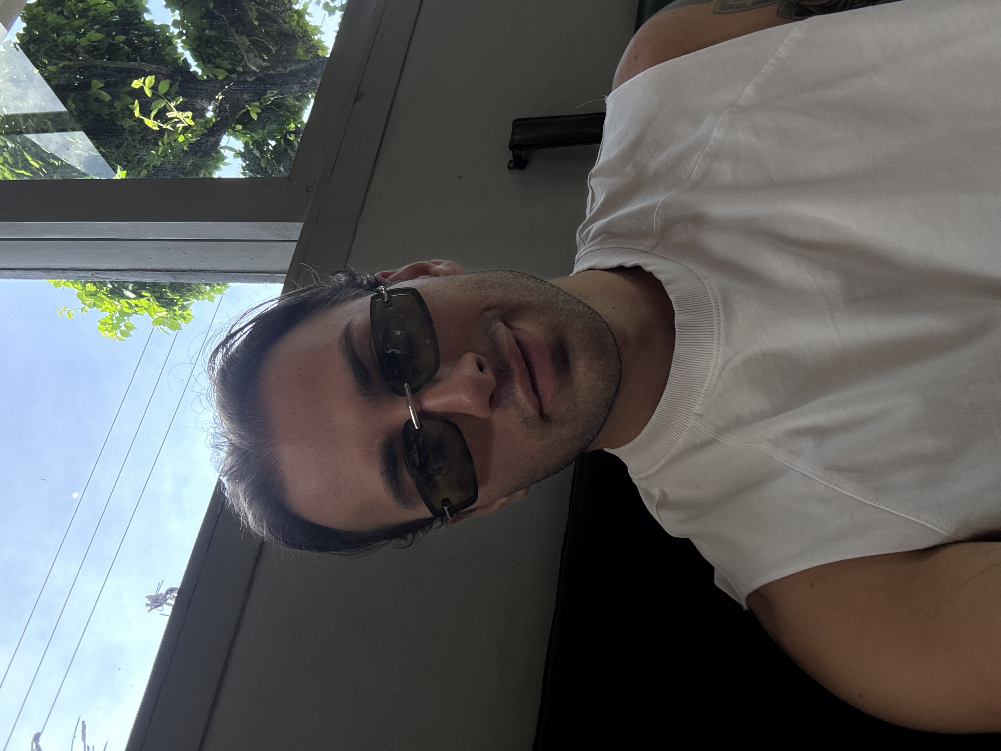 A medium shot shows a person with brown hair wearing sunglasses and a white t-shirt. They are sitting, with a black surface visible below them. Outside the window, trees and the sky are visible. The location is likely Mengwi, Indonesia, based on user information.