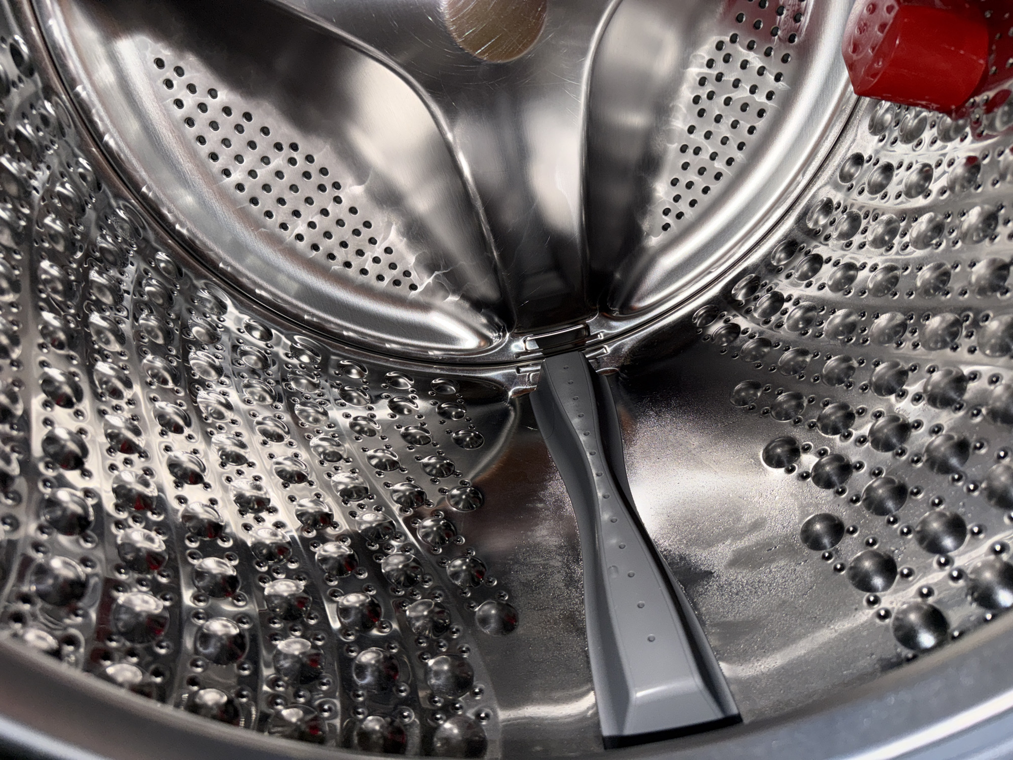 The image displays the interior of a washing machine drum. The drum is made of polished stainless steel and features numerous small perforations and raised dimples. A grey plastic component, likely a detergent dispenser or a part of the agitator mechanism, is visible on the left side of the drum. The surface of the drum appears slightly damp in some areas. In the lower right corner, a small, translucent red cube-shaped object is present. The lighting suggests an indoor environment, possibly a laundry room. No specific location in Varna, Bulgaria is discernible from the image itself.