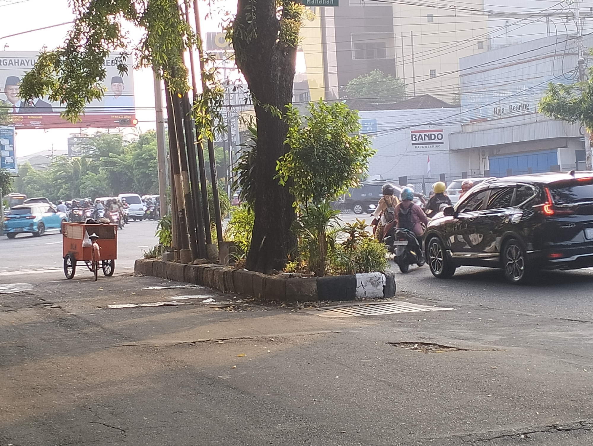 The image depicts a street scene in Surakarta, Indonesia. A person is pushing a red cart with wheels, possibly a street vendor. Several vehicles, including cars and motorcycles, are visible on the road. The environment appears to be an urban setting with buildings and trees lining the street. The sun is shining, casting shadows on the road. There are traffic lights and signs present, indicating a busy intersection. The overall impression is that of a typical day in a bustling Indonesian city.