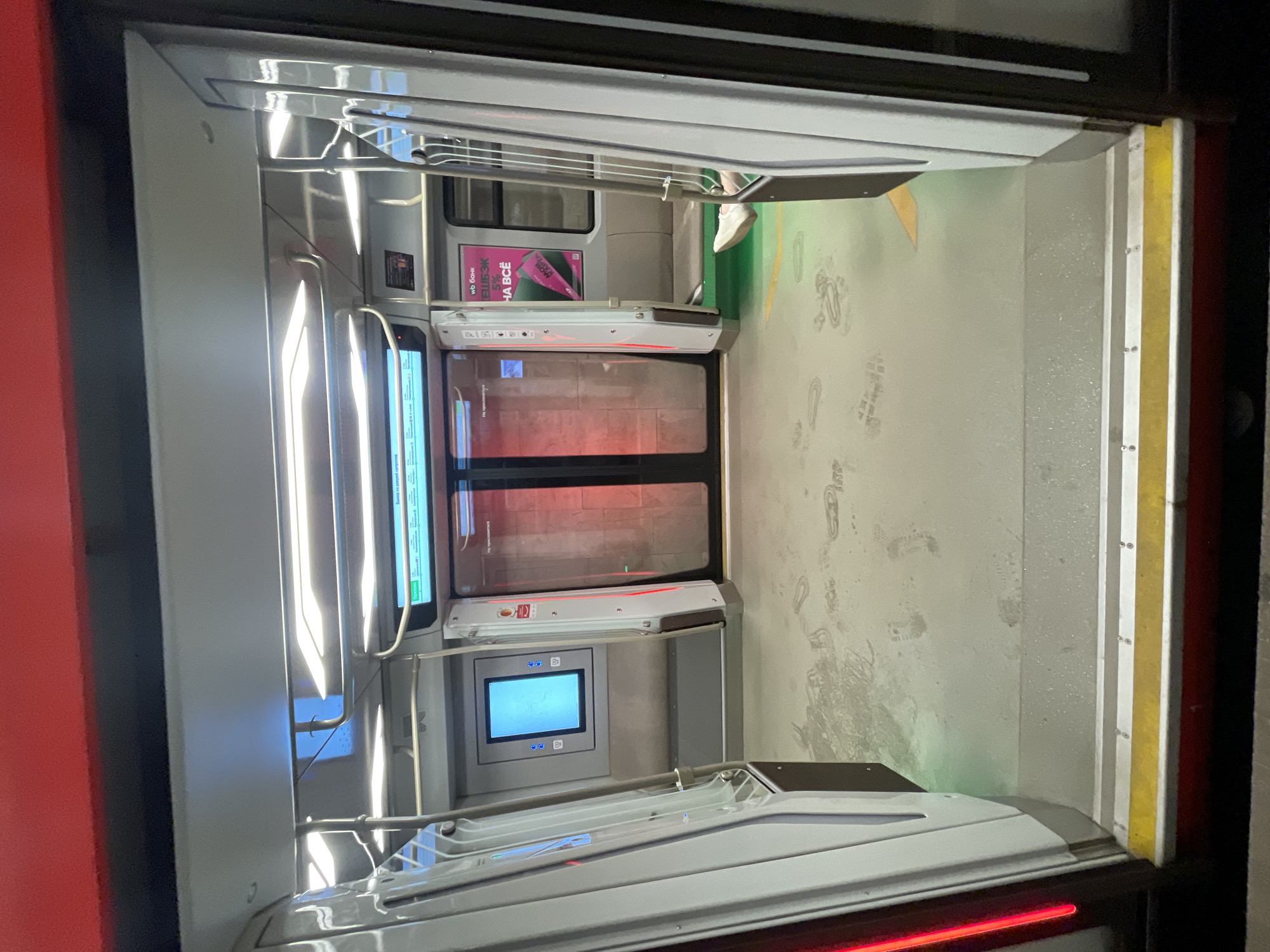 The image depicts the interior of a Moscow Metro train car in Russia. The scene is from within the train, looking through the open doors into the next car. The doors are partially visible, with electronic displays above and below the windows. The floor is light gray with numerous footprints. Yellow safety markings are along the edge of the floor. Metal handrails are visible on both sides of the train car. A poster with text and images is seen in the adjacent car. A person's foot in a white shoe is visible in the next car. The lighting is bright and primarily from the interior of the train car.