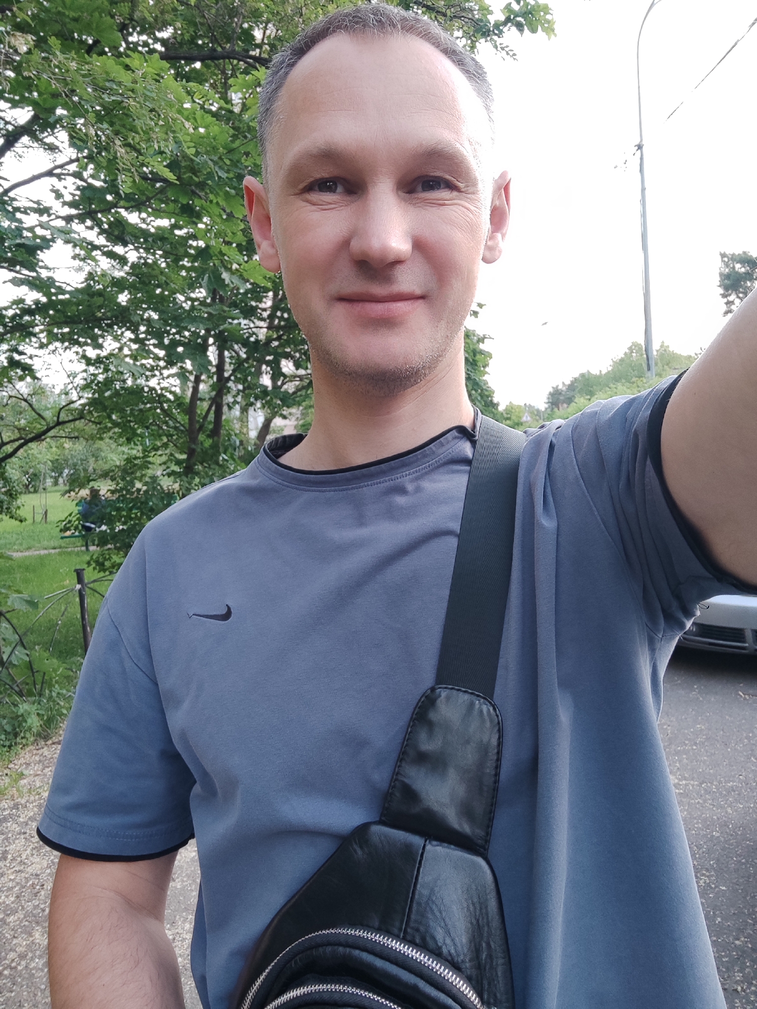 The scene is set outdoors in Kyiv, Ukraine, featuring a head-and-torso selfie of an adult male. The man, appearing to be in his late 30s to early 50s, has short, greying dark hair, a clean-shaven face, and light skin, displaying faint wrinkles around his eyes and a subtle smile. He is wearing a plain greyish-blue short-sleeved t-shirt with a black Nike swoosh logo on the left chest and black trim along the collar and sleeve cuffs. A black leather-like sling bag, featuring visible silver zippers, is worn across his chest from his left shoulder to his right hip. The man's right arm is partially visible on the right edge of the frame, indicating he is holding the camera.
The background consists of an outdoor environment with significant greenery. To the man's left, dense deciduous trees with green foliage are prominent, and a blurred figure of another individual appears to be seated or reclined in the mid-distance. To his right, a bright, possibly overcast sky is visible above a slender utility pole or street lamp. A light-colored car is partially visible on a paved surface behind the man's right arm and shoulder. The immediate foreground shows a lighter, textured paved path on the left and a darker paved surface on the right.