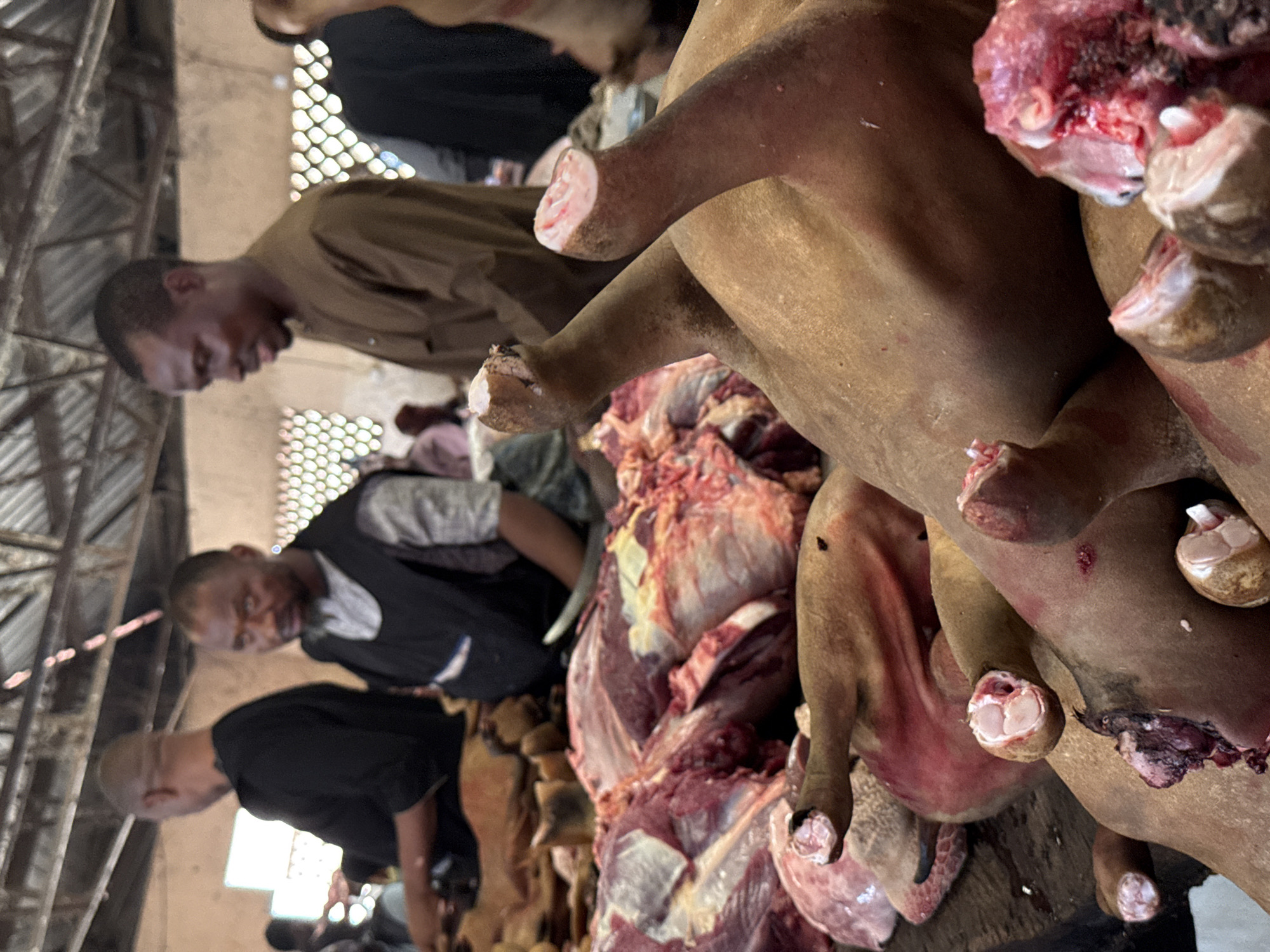 The image shows a butchering scene, likely in Lugbe, Nigeria, given the context. Three men are visible, one wearing a brown shirt, another a black shirt and the third a gray shirt. They appear to be working on large carcasses of meat, possibly beef or goat, with a significant amount of raw meat and severed limbs visible. A knife can be seen. The setting appears to be an open-air market or slaughterhouse, with sunlight filtering through a lattice-like structure.