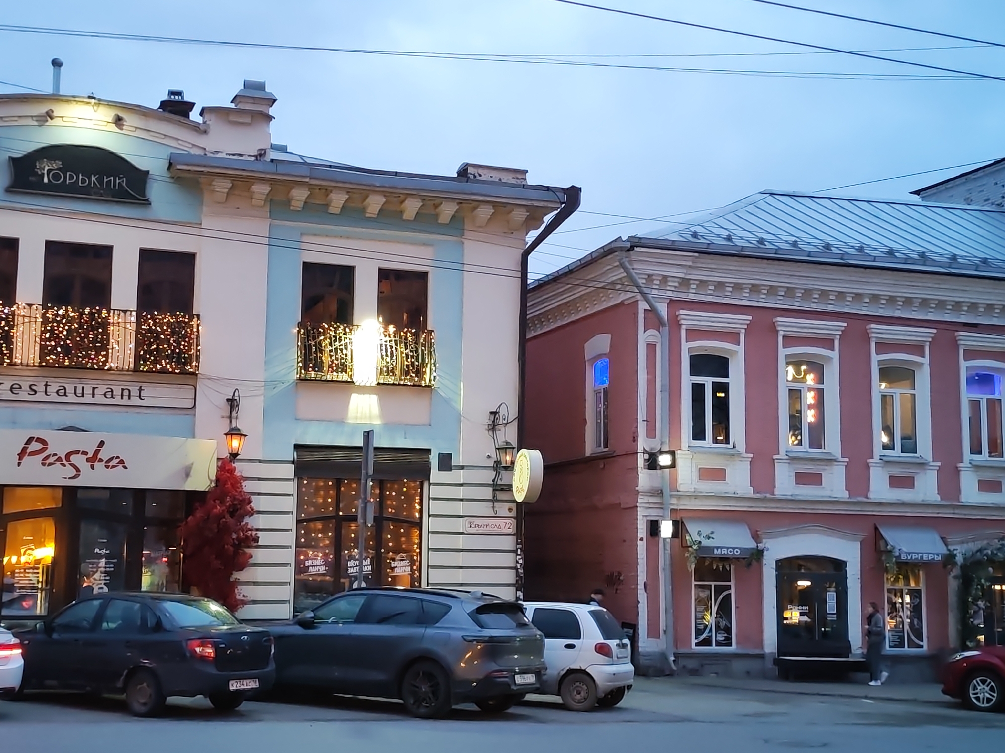 The image depicts a street scene in Izhevsk, Russia, at dusk or early evening, with artificial lights illuminating the buildings and shops. The sky is overcast, and power lines are visible overhead.
Two distinct multi-story buildings dominate the view. The building on the left features a light blue and white facade with two balconies decorated with warm string lights. A sign at the top reads "ГОРЬКИЙ" (GORKY). Below this, a "restaurant" sign is visible, with a prominent "Pasta" sign indicating a ground-floor eatery. Its windows display text such as "БИЗНЕС ЛАНЧИ" (BUSINESS LUNCHES) and "ВКУЛЫЕ ЗАВТРАКИ" (DELICIOUS BREAKFASTS). A small red decorative tree stands near the entrance. A street sign on this building reads "Крылова 72" (Krylova 72).
To the right, a reddish-pink building with white architectural trim houses several businesses. Awnings over its ground-floor shops display "МЯСО" (MEAT) and "БУРГЕРЫ" (BURGERS). The name "Ронни" (Ronny) is visible above an entrance below the "МЯСО" awning.
Several cars are parked along the street: a dark grey SUV, a black sedan with license plate "К 234 АС", a white compact car, and a red car partially visible on the far right. A few pedestrians are seen on the sidewalk. The scene conveys a quiet urban atmosphere as businesses light up for the evening.