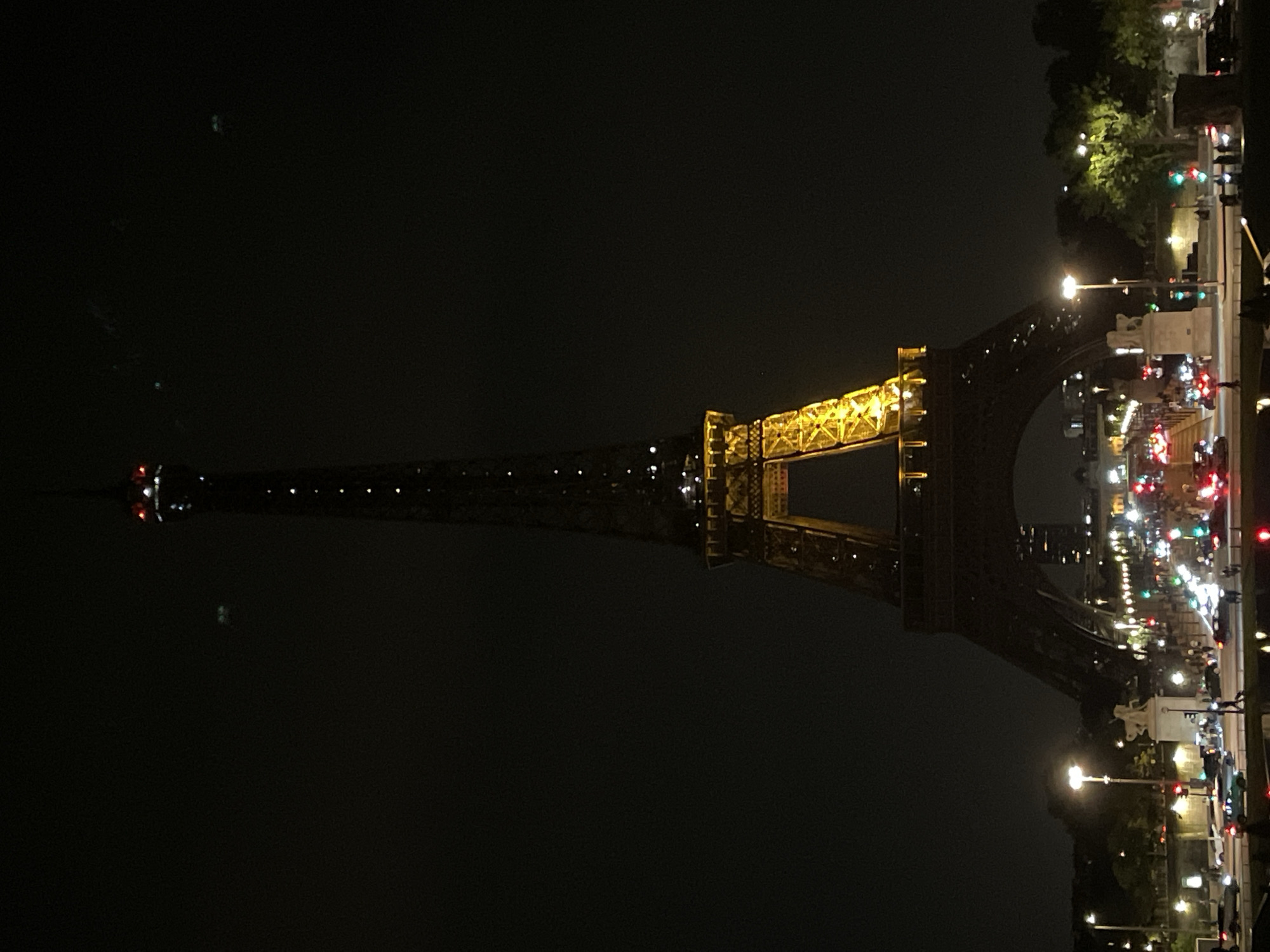 The image shows a night-time view of the Eiffel Tower in Paris, France. The tower is illuminated with golden lights, and the top is visible against a dark sky. In the foreground, there is a street scene with traffic, streetlights, and some pedestrians. The lights of the city are visible in the distance.