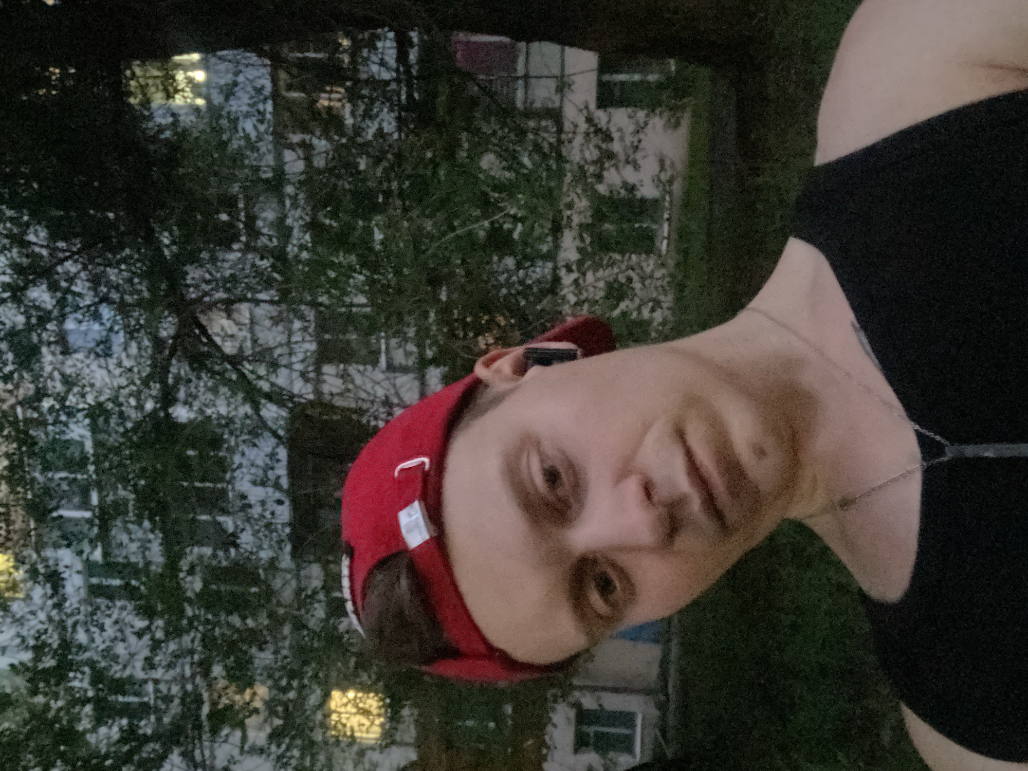 A young man wearing a red baseball cap and a black tank top is pictured outdoors in front of a building. The man has brown hair and is looking towards the camera. He is wearing a silver necklace and earbuds. The building appears to be an apartment building with visible windows, some of which are lit. Trees and grass are visible in the foreground and background. The overall lighting suggests it is dusk or evening. The provided context indicates this scene is located in Samara, Russia.
