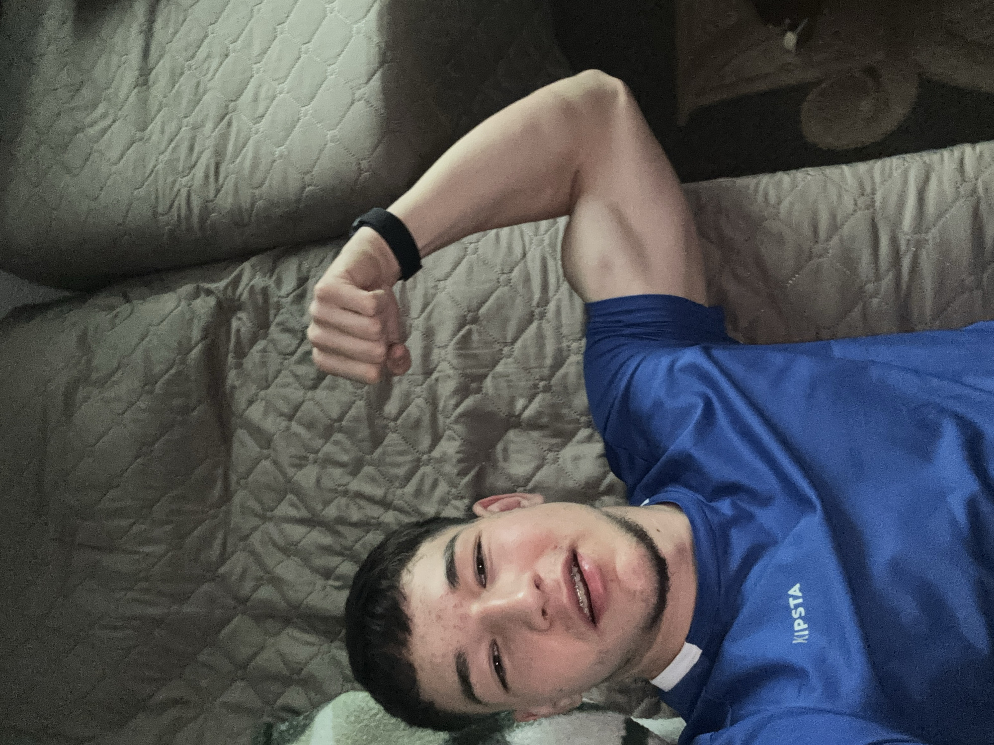 A person wearing a blue sports shirt with the brand "KIPSTA" visible is lying on a quilted, neutral-colored bed. The person has short dark hair, visible braces on their teeth, and a light beard. Their left arm is flexed upwards, displaying a black wristband. The background is a dimly lit room. This image does not contain any details about the city of Zlatograd, Bulgaria.