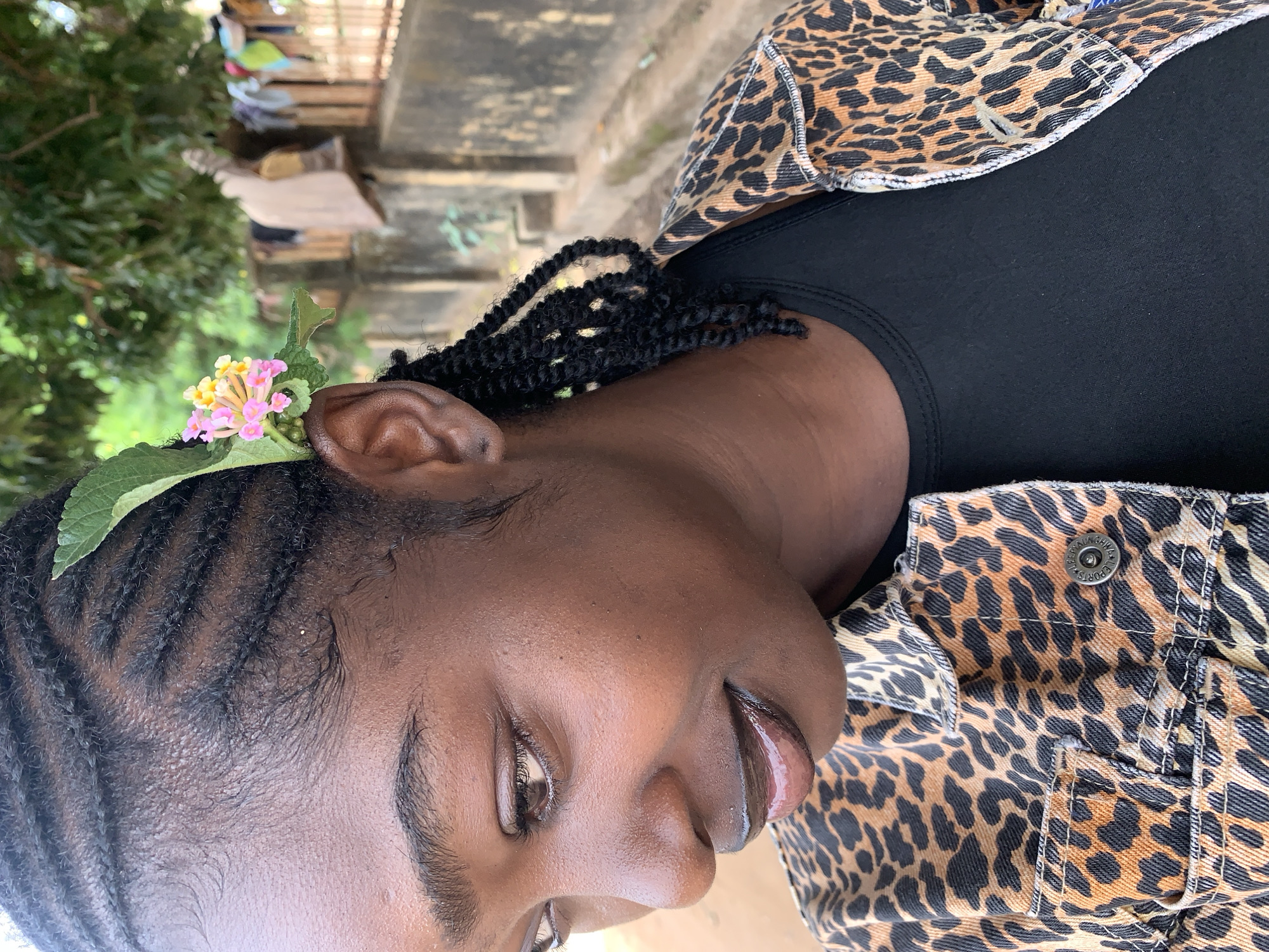 An outdoor, eye-level, medium close-up shot features a young adult female of Black African descent, positioned slightly left of center and turned towards the left, looking downwards. Her expression is calm with a slight smile. She has dark skin, with visible pores and a subtle sheen. Her eyes are dark, and her full lips are covered in a dark, glossy product. Her hair is styled in cornrows leading into twisted tendrils that fall behind her right ear. Tucked into her hair above her right ear are small, delicate pink and yellow flowers accompanied by green leaves. She is wearing a black crew-neck t-shirt beneath a denim jacket with a prominent leopard print pattern (orange-tan with black spots). A silver-colored metal button with circular text, "I SLEEP AND WAKE UP," is visible on the right side of her jacket. The background indicates an outdoor setting in what appears to be a residential or semi-urban area. To the right, a weathered, dark grey concrete or stone wall runs horizontally, behind which is a fence made of vertical wooden slats or bamboo. Clothes are visible hanging to dry behind this fence. Green foliage and tree branches are present in the upper and mid-background. The ground in the lower left appears to be an unpaved surface. This scene is situated in the city of Rafin Par, Nigeria.