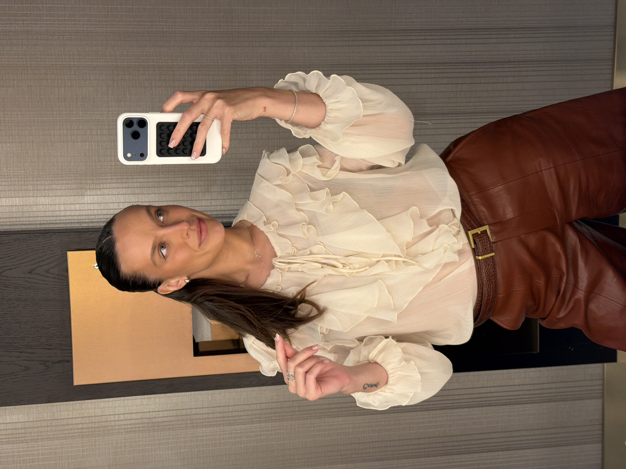 A woman is taking a selfie in a room with a textured gray wall and a dark wood panel with a copper accent. She is wearing a cream-colored ruffled blouse and brown leather pants with a patterned belt. She has her hair pulled back in a ponytail and is wearing earrings, a necklace, rings, and a bracelet. She is holding a white phone in her right hand. The image does not provide any information about the location being in Budva, Montenegro.