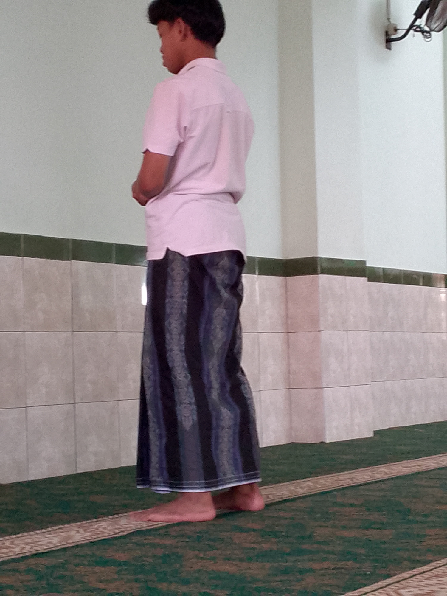 A young person with dark hair stands with their back to the camera, facing a wall. They are wearing a light pink short-sleeved shirt and a dark blue and black striped sarong. The sarong is patterned with vertical stripes that vary in width and color intensity. The person's feet are bare and resting on a green carpet with a patterned border. The background consists of a pale green wall above a tiled section. The tiles are rectangular and light-colored, arranged in horizontal rows. A dark green tile band separates the wall from the tiled section. A light fixture is visible on the upper right side of the wall. The scene appears to be indoors, likely in a place of worship or a communal area, given the attire and the carpet. The lighting suggests an ambient indoor environment. The overall impression is one of quiet contemplation or waiting. The location is identified as Cimahi, Indonesia.