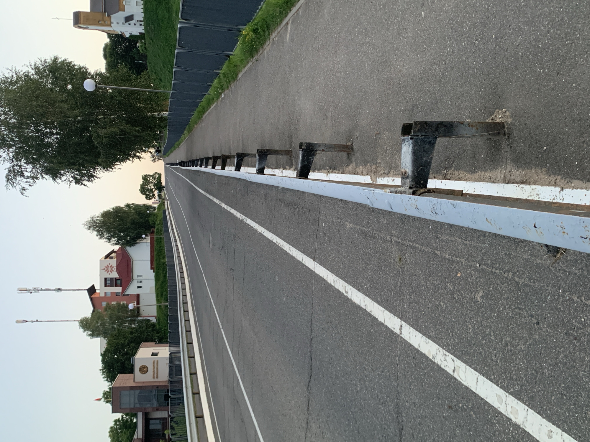 The image depicts a road in Kapyĺ, Belarus, with a sidewalk adjacent to it. The road is asphalt and has a white line dividing the lanes. A pedestrian walkway with a metal guardrail runs along the right side of the road. In the background, there are buildings and trees. One building appears to be a multi-story structure with antennas on top, and another is a smaller white building with a red roof. The sky is clear and appears to be at dusk or dawn. There are no people visible in the image.