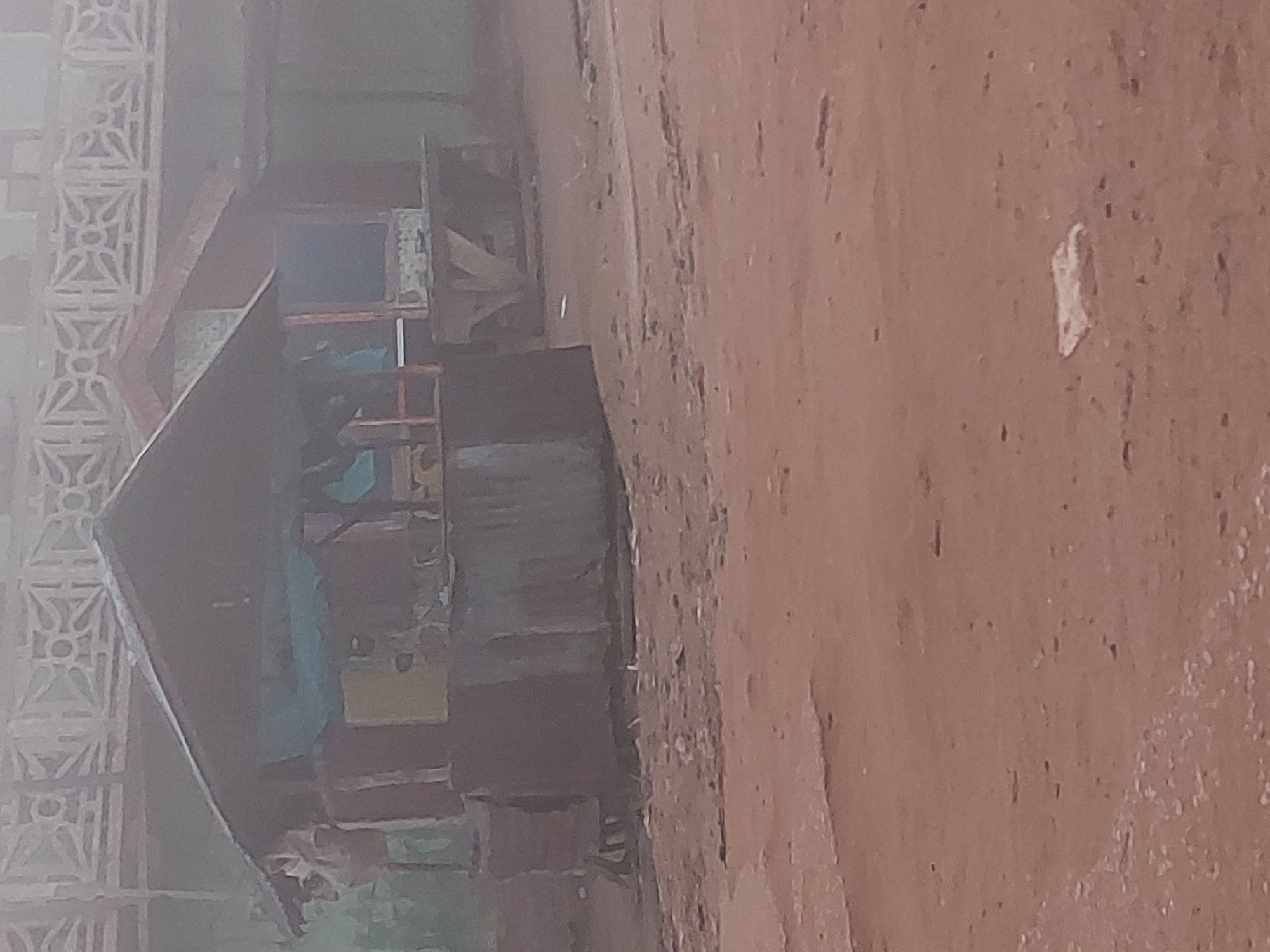 The image depicts an outdoor scene with two makeshift stalls or small structures constructed from wood and corrugated metal. The stalls are situated on a dirt ground. The stall on the left is partially obscured by a blue tarp and has a dark roof. The stall on the right is less visible and appears to have a lighter-colored structure behind it. In the background, there is a decorative concrete wall or fence with geometric patterns. The overall atmosphere is hazy, suggesting either fog or dust, which lends a muted quality to the colors and details of the scene. The image is undated and uncaptioned, making it difficult to pinpoint the exact time and day it was taken. The scene gives the impression of a market or a roadside area in a less developed urban setting, potentially in Bukuru, Nigeria, given the prompt's reference. There are no people visible in the image, and no specific actions are being taken. The environment appears dry and dusty.