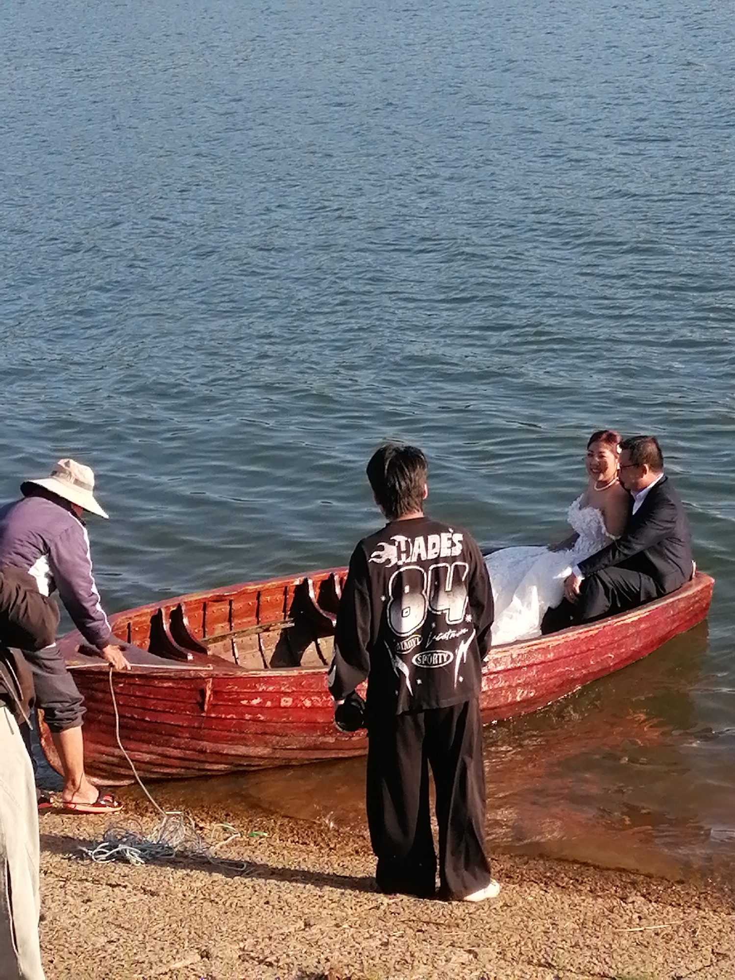 A wedding photoshoot is underway on a small, red wooden rowboat near the shore of Xuan Huong Lake in Da Lat, Vietnam. A bride in a white wedding gown and a groom in a dark suit are seated inside the boat, facing the camera. A man wearing a black shirt with graphics and a woman in a wide-brimmed hat and casual clothing are also present near the boat. The man in the hat appears to be securing the boat to the shore with a rope. The environment is a body of water, likely a lake, with calm ripples. The shore is covered in pebbles. The scene suggests a posed photograph being taken in a natural, outdoor setting.