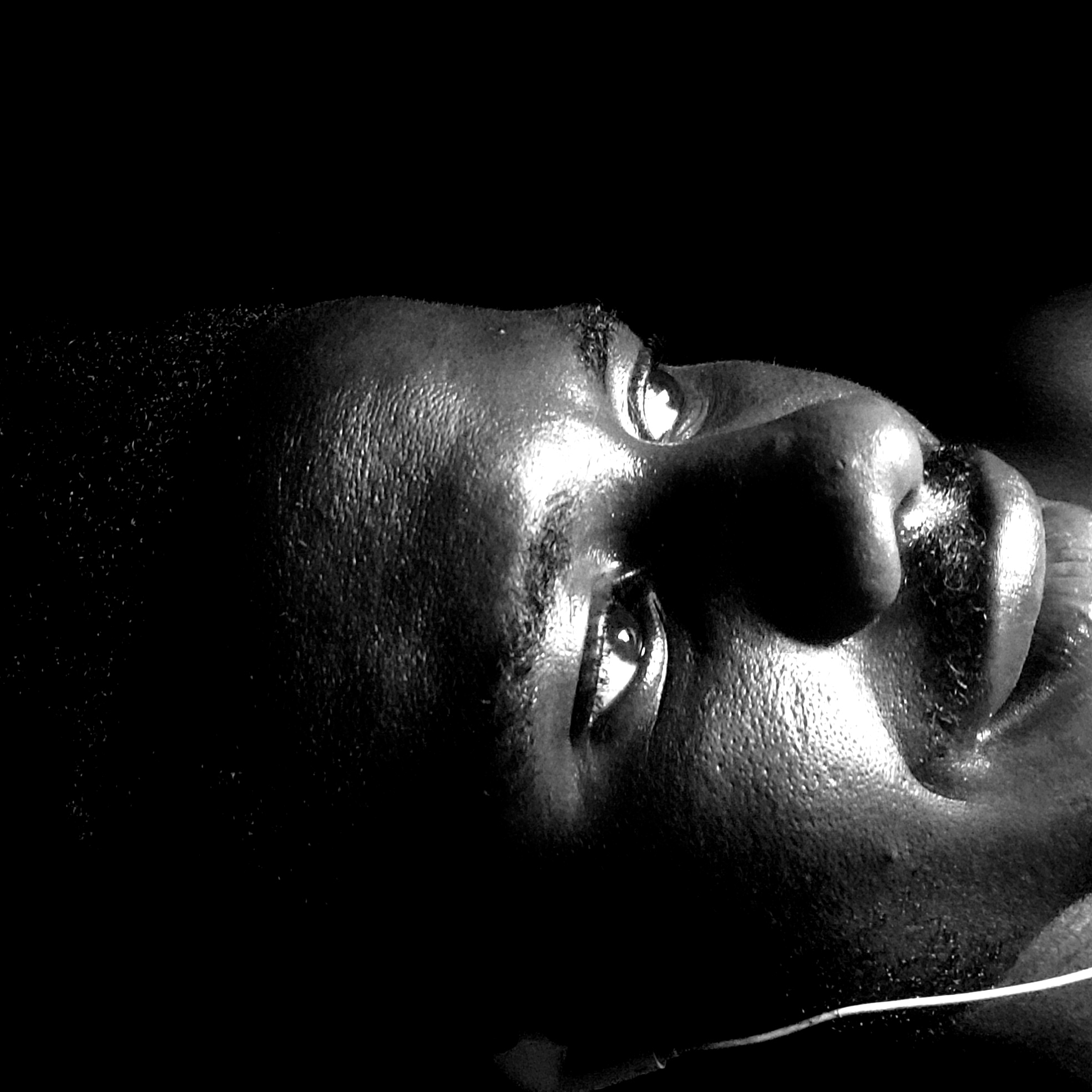 A close-up, black and white photograph depicts a man's face in stark lighting. He is lying on his back, looking upwards. The man has dark skin, short hair, and facial hair around his mouth. A thin, white wire or string is visible across his lower face, possibly an earphone cord. The location of Narayi, Nigeria, is not directly discernible from the image itself. There are no other contextual clues in the image to indicate location beyond the subject's features.