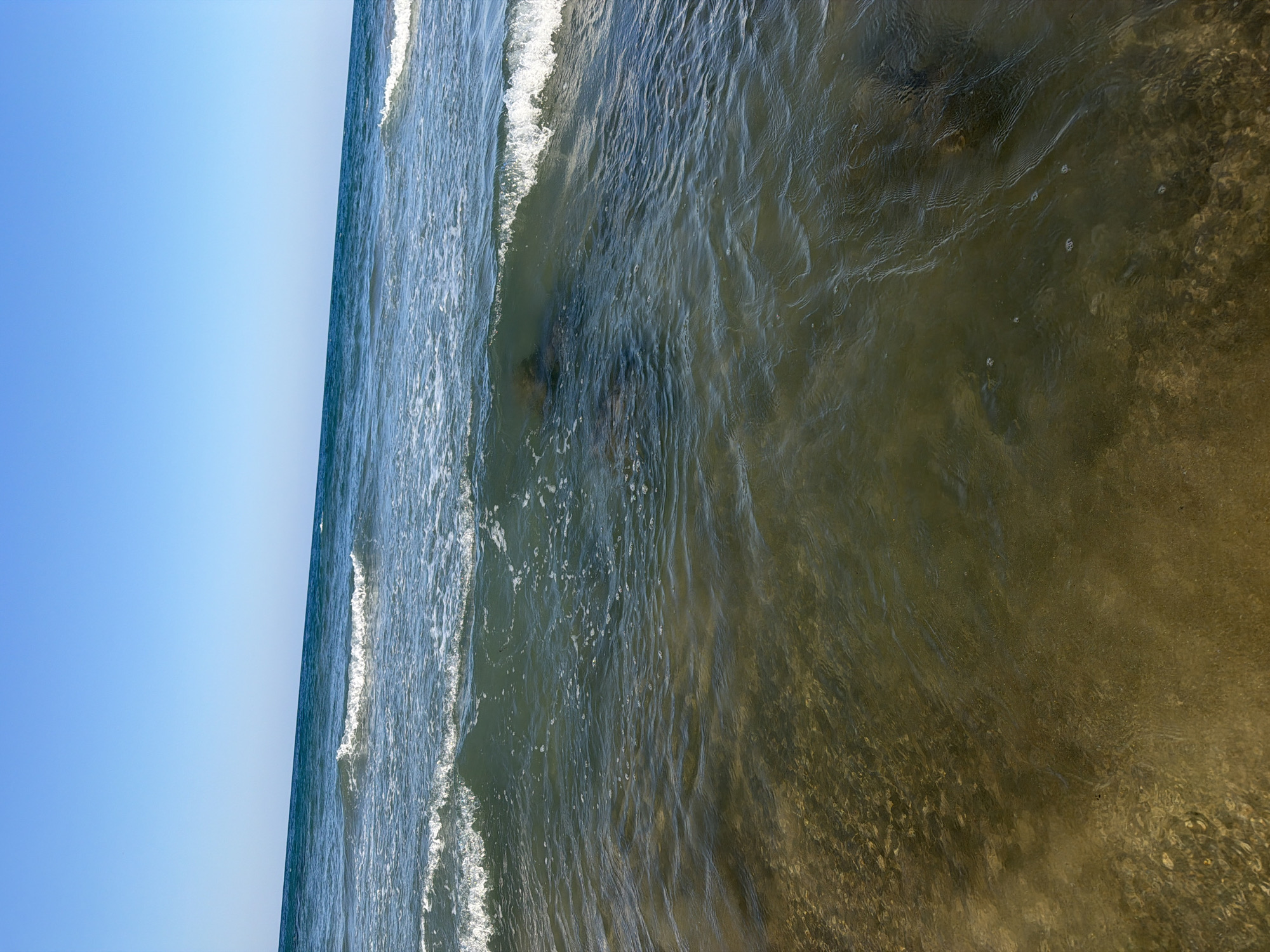 The image depicts a beach scene in Georgioupoli, Greece. The foreground features shallow, clear water revealing a sandy and pebbly seabed. Gentle waves are breaking, with white foam washing ashore. In the mid-ground, darker shapes are visible beneath the water's surface, possibly rocks or submerged objects. The horizon is marked by the meeting of the dark blue sea and a bright, clear blue sky, with no landmass visible. The overall impression is one of a calm, sunny day at the coast.