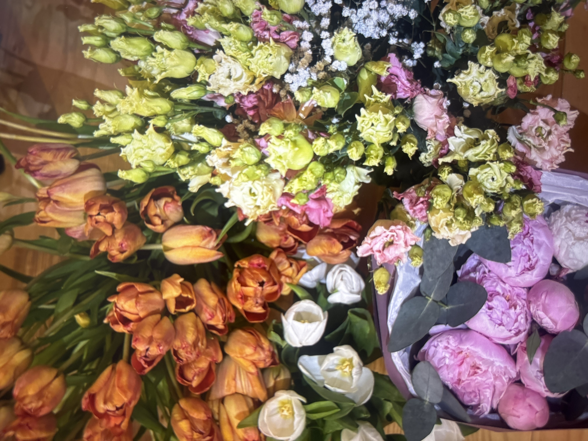A top-down shot reveals a dense collection of flowers. Predominantly, orange tulips with green foliage stand out, arranged alongside yellow and white flowers. Mixed within are smaller white flowers resembling baby's breath, and pink peonies arranged in a compact bunch. The arrangement is casually placed on a wooden surface, the specific location and city remain unknown.