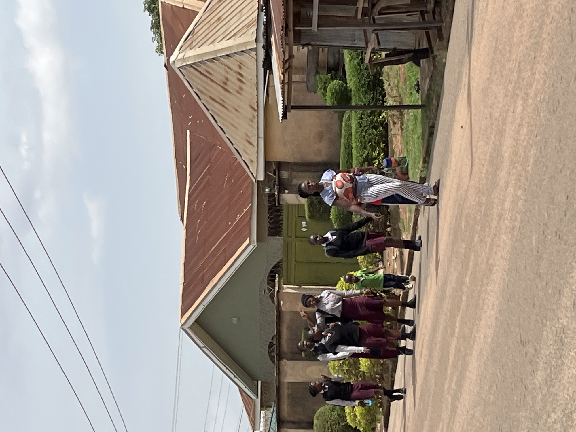 In Jos, Nigeria, a group of schoolgirls in maroon skirts and white shirts, some wearing black blazers, stand in a line in front of a building with a brown and grey metal roof. To the side is a woman holding a soccer ball. They are on a paved area near a green building.