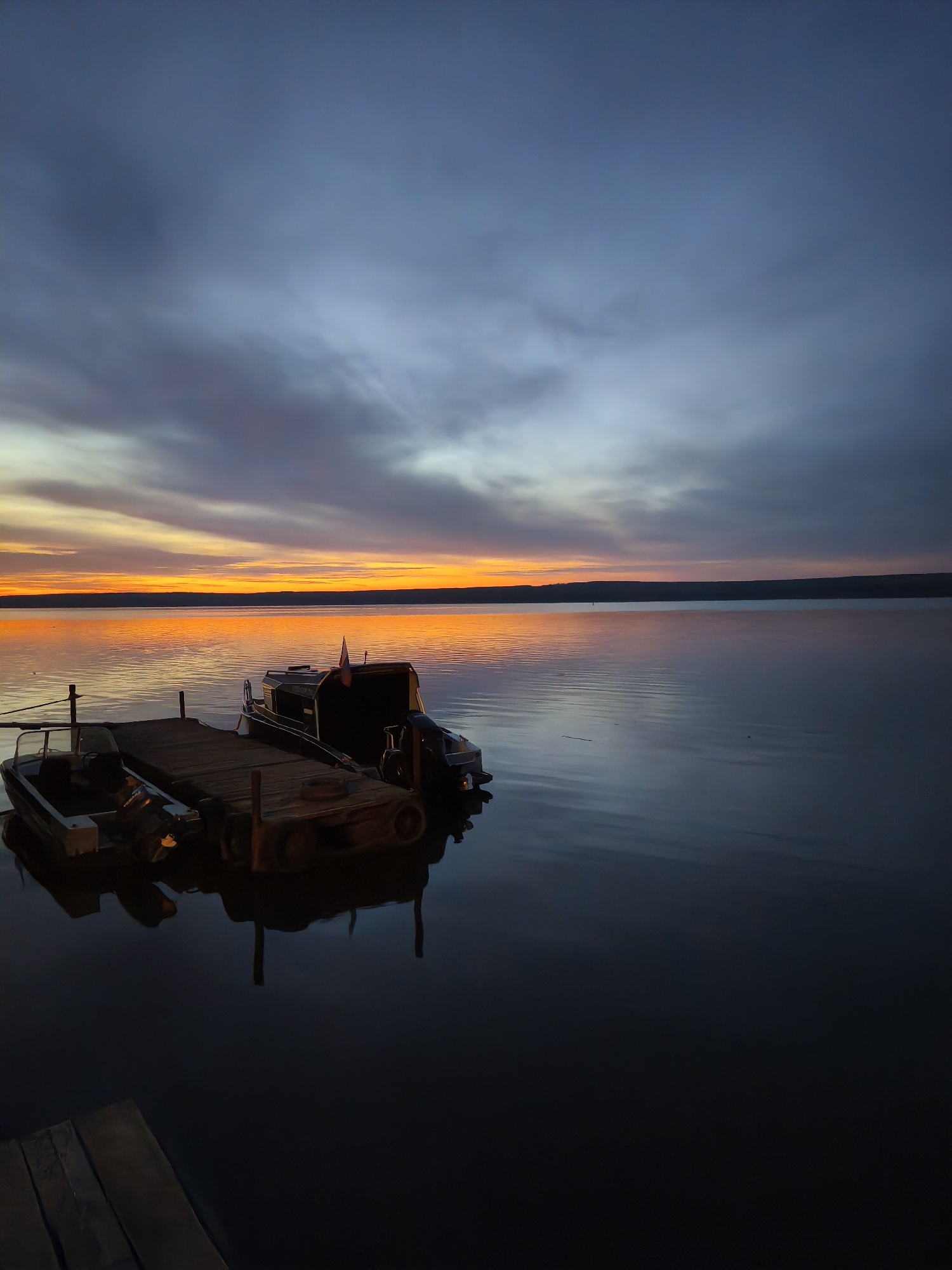 The image shows a serene evening scene at a lake, presumably the Kama Reservoir, near the city of Chaikovsky, Perm Krai, Russia, given the provided context. A small wooden dock extends into the calm water, with two boats moored alongside. The boat closer to the viewer appears to be a smaller, open-bowed vessel. Adjacent to it is a larger boat, potentially a cabin cruiser, equipped with an outboard motor. The sky displays a gradient of colors ranging from dark blue and gray at the top to vibrant orange and yellow hues at the horizon, reflecting on the still water. A treeline is visible in the distance, marking the far shore of the reservoir. The light suggests that this image was captured at twilight or sunrise.