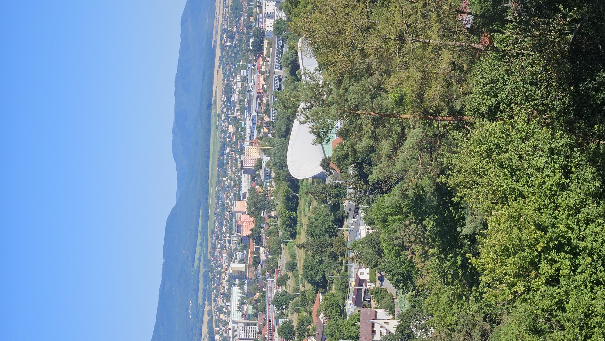 The aerial image captures a panoramic view of the city of Prešov, Slovakia, on a clear, sunny day. The foreground is dominated by lush green trees, with dense foliage and tall pines creating a natural frame. In the middle ground, a sprawling urban landscape unfolds, characterized by a mix of residential areas with red-tiled roofs and apartment buildings, as well as some larger structures. Prominent among these is a distinctive white, curved roof building, likely an arena or sports complex, situated amidst the greenery. Beyond the city, a range of rolling blue-hued hills and mountains forms a distant backdrop under a vast, cloudless blue sky. The scene suggests a city integrated with its natural surroundings, offering a view from an elevated vantage point.