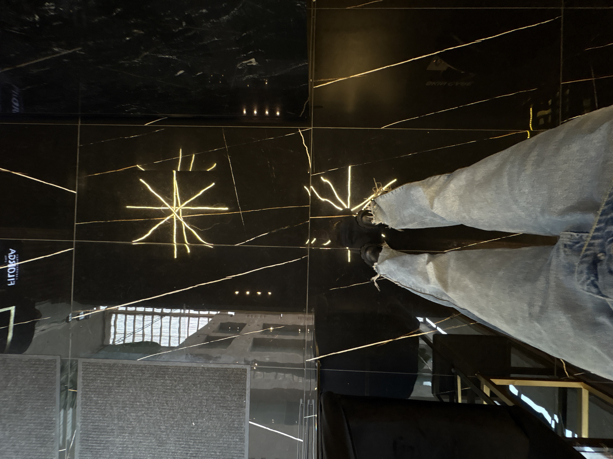 The image shows a person wearing blue jeans standing on a glossy black tiled floor with gold-colored veins or patterns. The floor reflects the surroundings, including a building with visible windows and a textured surface. The person's feet and shoes are visible, and part of a black chair is in the lower part of the frame. Based on metadata, the location is Ciudad López Mateos, Mexico.