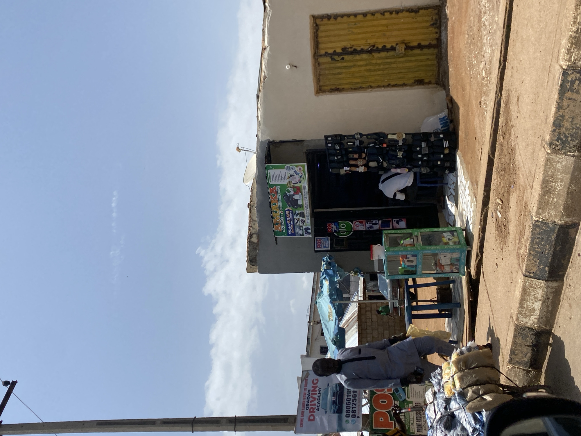 This media file depicts a street scene in Gura Top, Nigeria, likely during the daytime given the bright sunlight casting shadows. The image is oriented vertically.
The primary subject is a shopfront selling various items, including what appear to be shoes displayed on shelves and a display case with mobile phones and accessories. A person in a light gray garment is seated next to a stack of bundled goods, possibly merchandise, on a cart. Another person, partially visible, is standing inside the shop.
Several signs are visible. One prominently advertises "GRADAN DRIVING ACADEMY" with contact numbers. Another sign for the shop reads "AMAKA ACCESSORIES COMMUNICATIONS" and "Victor BRAVE SHOES COLLECTION'S," along with a phone number. There are also signs with "POS" and "glo," suggesting telecommunication or financial services.
The setting is an urban environment with a weathered building featuring a yellow corrugated metal door. The street itself appears to be paved with rough concrete and bordered by a curb. The sky is visible with some clouds, indicating fair weather. The overall impression is of a bustling street market or commercial area.