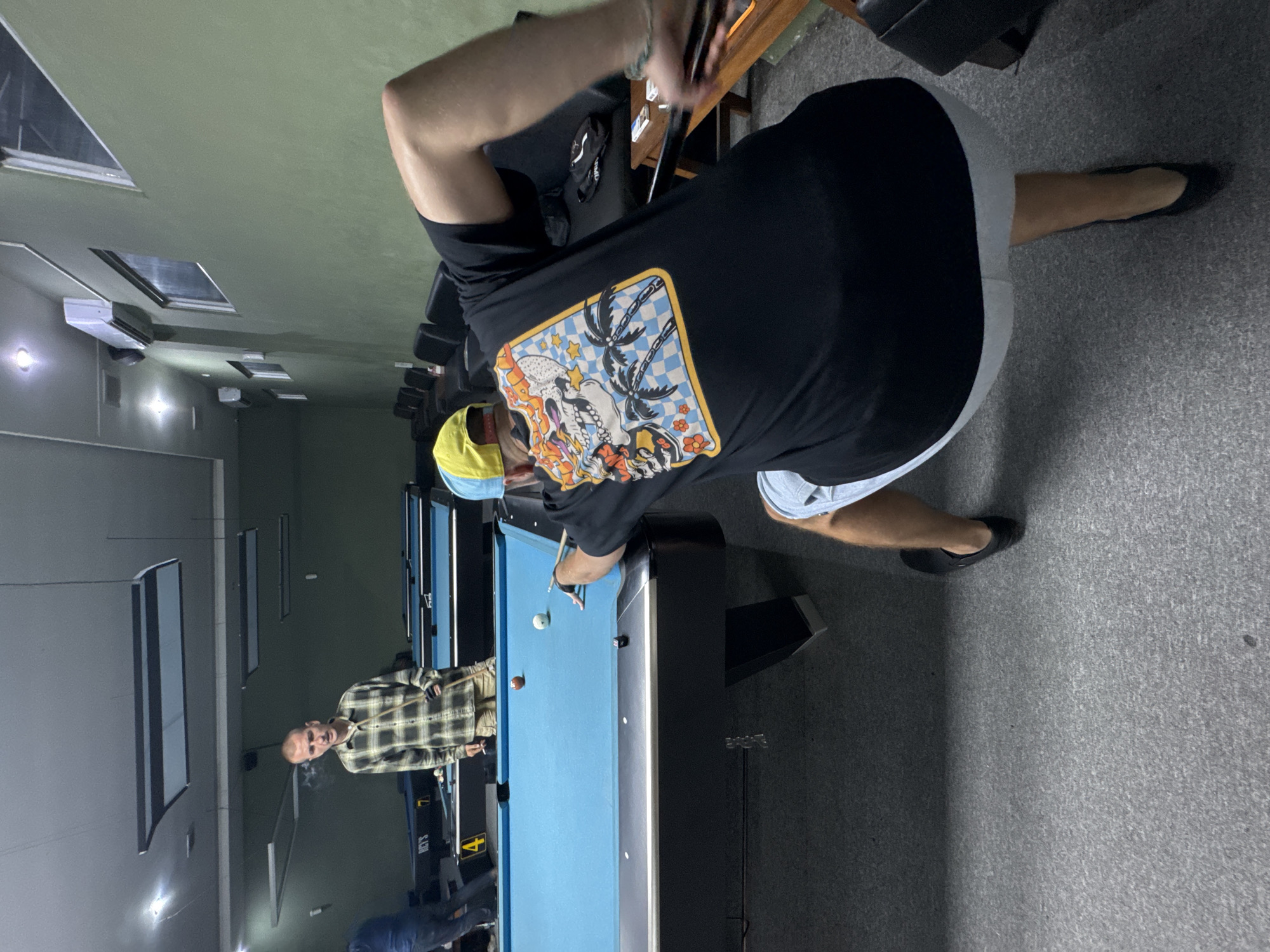 The image depicts an indoor pool hall in Kuta Selatan, Indonesia. Two men are actively engaged in playing pool. The man in the foreground, wearing a black t-shirt with a skull graphic on the back and a yellow and blue baseball cap, is in the process of taking a shot, his body low and focused on the game. The other man, standing in the background and wearing a plaid shirt and shorts, appears to be observing the shot, with a cigarette in his mouth. Multiple pool tables are visible in the dimly lit environment, with blue felt surfaces and dark wood trim. Chairs are arranged along the walls, and overhead lighting fixtures illuminate the scene. The overall atmosphere suggests a casual recreational setting.