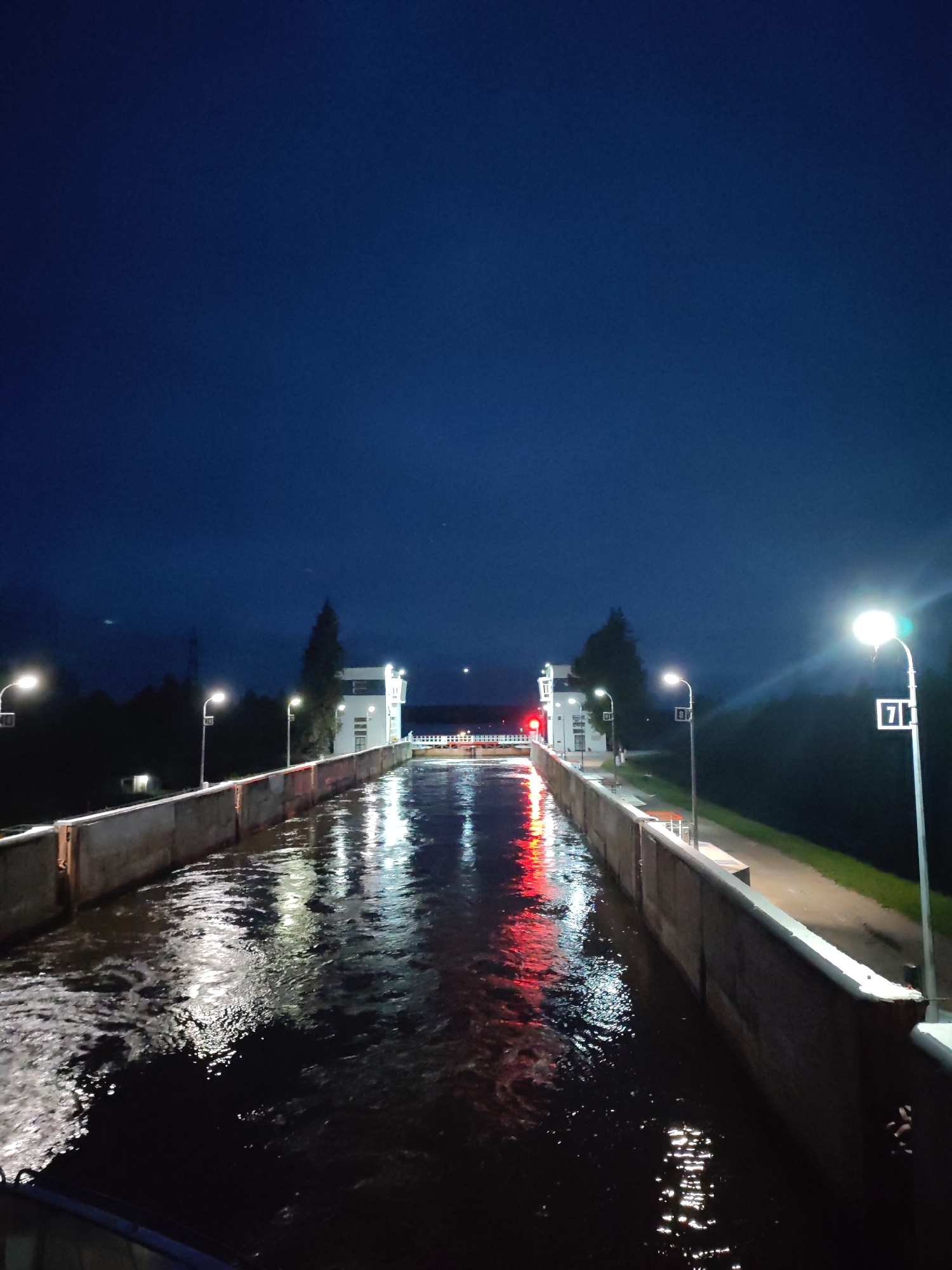 The image depicts a modern lock gate structure at night, likely part of the infrastructure of the city of Belousovo, Russia. The perspective is from a slightly elevated position, looking down a rectangular canal that is illuminated by artificial lights. The water within the canal is dark and reflects the lights from the surrounding structures, creating streaks of white and red.
On the sides of the canal are concrete walls, topped with railings. Along the right wall, there are several lampposts, each with a bright, white light source. One lamppost on the right is notable for a black sign with a white number '71' on it. Further along the right side, a paved walkway or path is visible, bordering a dark, tree-lined embankment.
On the left side of the canal, a similar concrete wall is present. In the distance, at the end of the canal, are two white, rectangular buildings. These buildings appear to be part of the lock gate mechanism, with lights visible on their facades. A red light is also visible on one of these structures, possibly indicating a warning or status. Trees are visible behind these buildings and along the left embankment, their silhouettes dark against the twilight sky.
The sky above is a deep, dark blue, characteristic of nighttime, with no visible stars or moon. The overall impression is one of functional industrial infrastructure illuminated for nighttime operation, situated within a natural or semi-natural landscape.