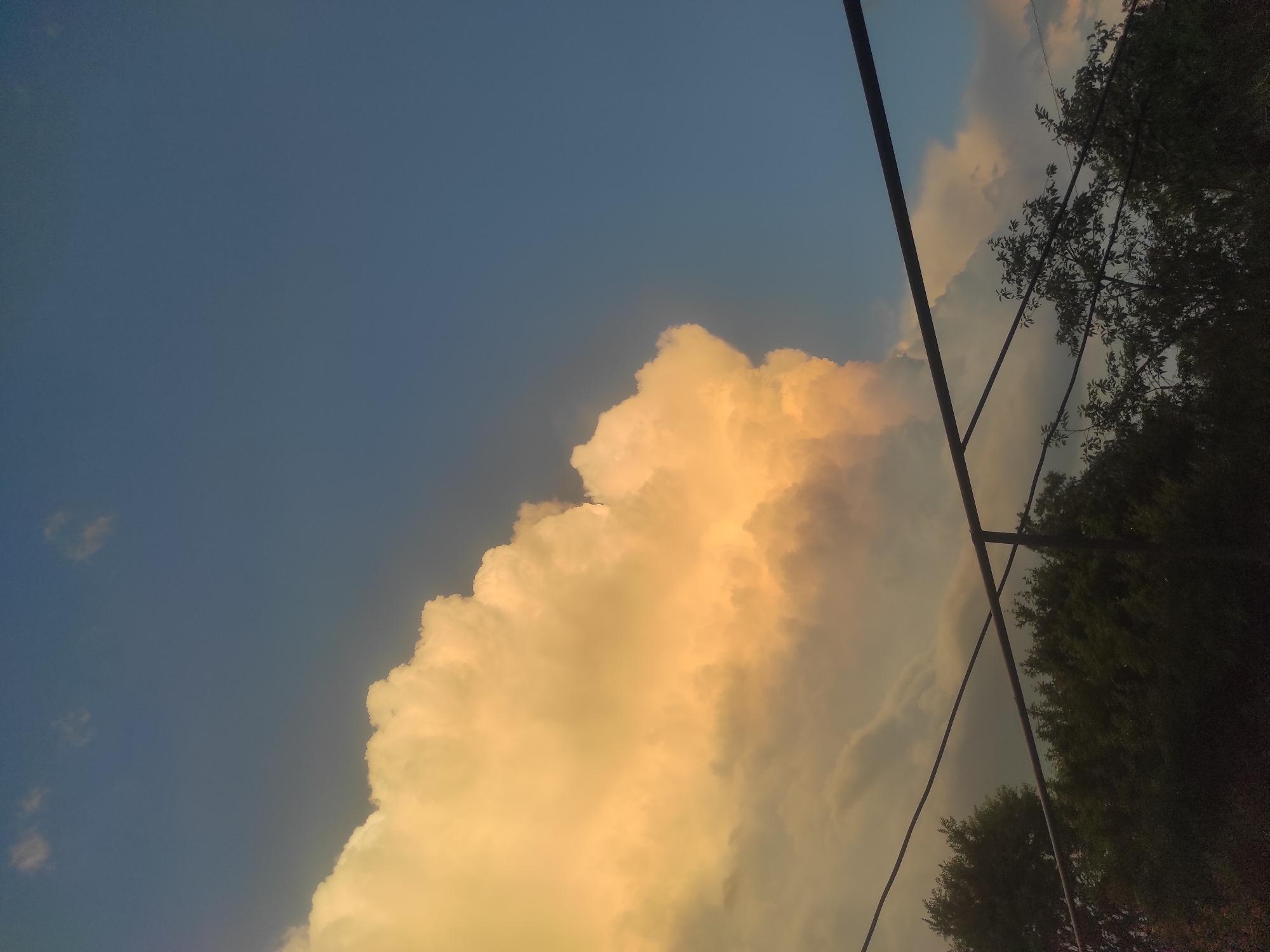 The image captures a scene from the border area of Gyumri-Akhuryan, Armenia. The sky dominates the upper portion of the frame, displaying a gradient of deep blue transitioning to lighter shades. A large, dramatic cumulus cloud, illuminated by a warm, golden light, occupies the left half of the sky. The lower part of the image is obscured by the dark silhouettes of trees and two parallel, dark horizontal lines, likely wires or pipes, stretching across the frame. The overall atmosphere suggests a late afternoon or early evening setting, possibly before or after a storm.