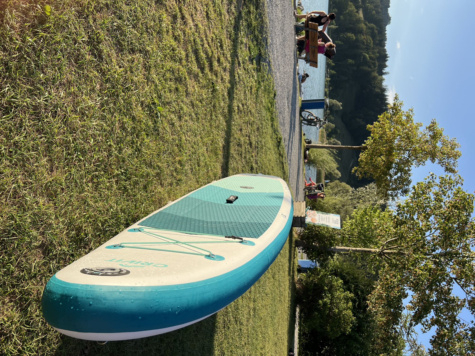 The image depicts a sunny day at the Alpsee lake in Immenstadt im Allgäu, Germany. In the foreground, a stand-up paddleboard lies on the grass. In the midground, several people are relaxing by the lake. Two women are seated on a bench near the water's edge, while other individuals are sitting or lying on the grass and shore. A bicycle is parked next to a blue trash bin. The background features a lush green forest and a clear blue sky. The scene suggests a leisurely outdoor activity at a popular lakeside spot.