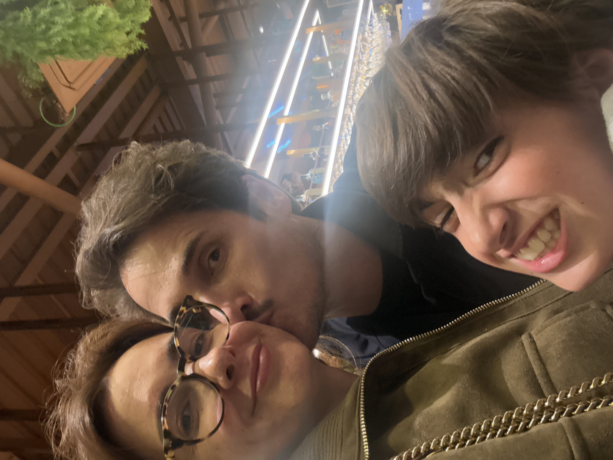 Three people are in the frame. The woman at the bottom left wears glasses and has short brown hair. A man with dark hair kisses her cheek. A child with short brown hair smiles to the right of the man. The individuals are indoors under a wood ceiling. The background has vertical lights and decorations. The location is Vila Nova de Gaia, Portugal.