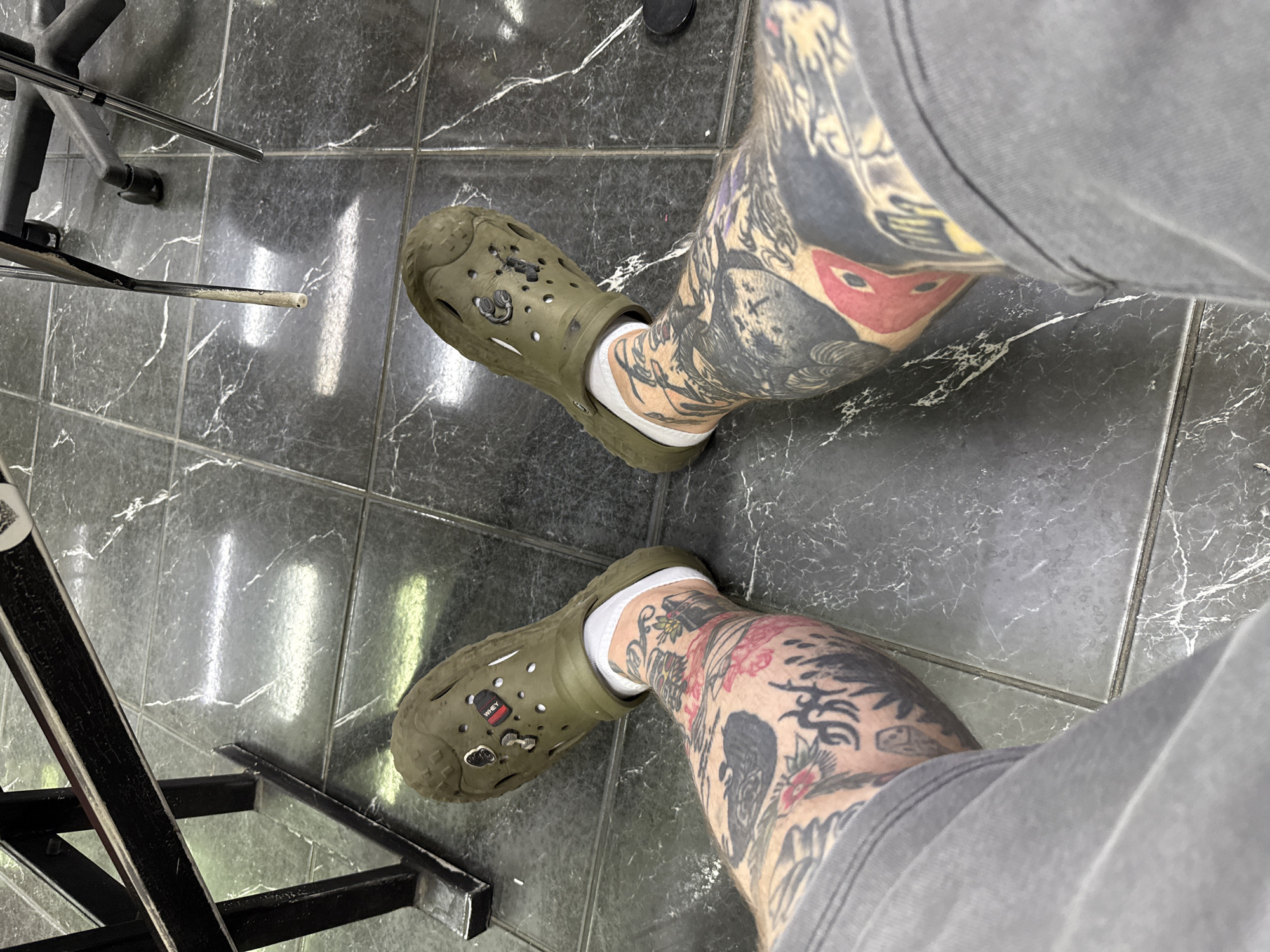 The image shows a close-up, top-down view of a person's legs wearing olive green Crocs and white socks. The legs are heavily tattooed with black and red ink. The Crocs have several Jibbitz charms, including one that resembles a dumbbell and another with the word "WHEY" on a black background. The person is sitting on a floor tiled with dark gray, marbled tiles. To the left and in the background, a black metal structure, possibly part of a table or chair, is visible. The overall setting appears to be indoors. There is no indication of the location being in Moscow, Russia, or any other specific city.