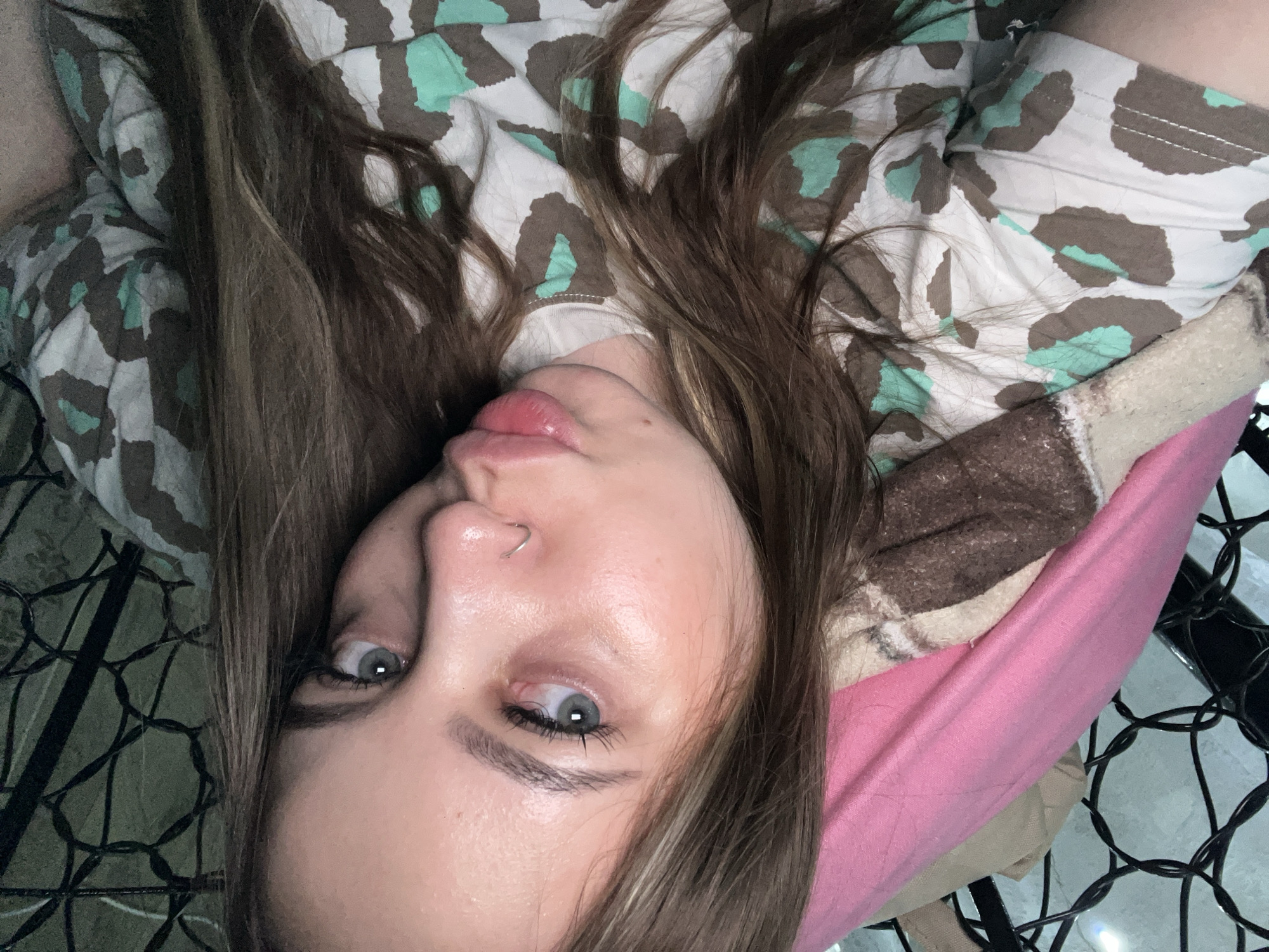 A close-up selfie shows a young woman with long brown hair. She has fair skin, light-colored eyes, and a nose ring. She is wearing a white shirt with a green and brown animal print. She appears to be lying down on a pink pillow with a brown and white patterned blanket nearby. Part of a black metal structure, possibly a chair or bed frame, is visible in the lower right corner. The location is not discernible from the image.