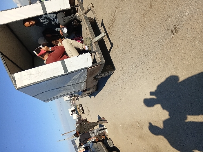 People sit inside a small cart with a white canvas top. One person is reading a book, while others are seated and looking around. The cart is on a paved road with other vehicles and people nearby. The shadow of a person taking the photograph is visible on the right side of the image. The sky is blue, and the lighting suggests it is daytime.
