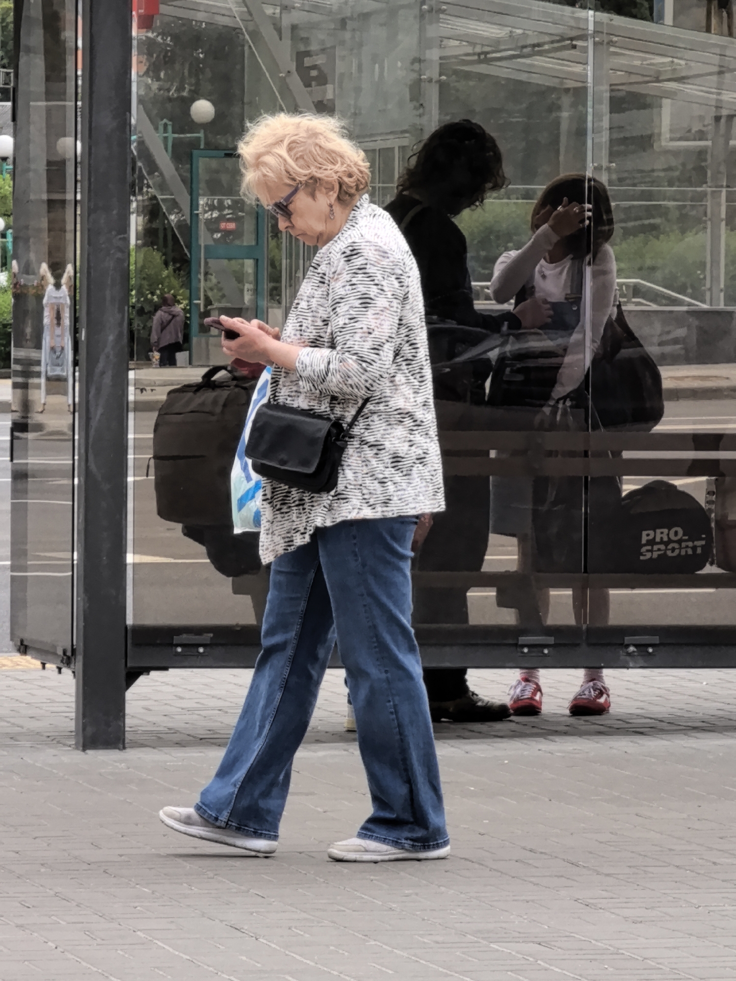 An elderly woman with blonde hair and sunglasses is walking on a paved sidewalk and looking at her smartphone. She is wearing a black and white patterned blouse, blue jeans, and white sneakers. A black purse is slung across her body. Behind her, a glass bus shelter is visible, with reflections of people and trees. Two younger individuals, their faces obscured, are seated inside the bus shelter, and a dark backpack is on the ground next to them. In the reflection of the glass, a person in a dark jacket is visible standing in the background. The overall environment appears to be an urban setting, likely a bus stop.