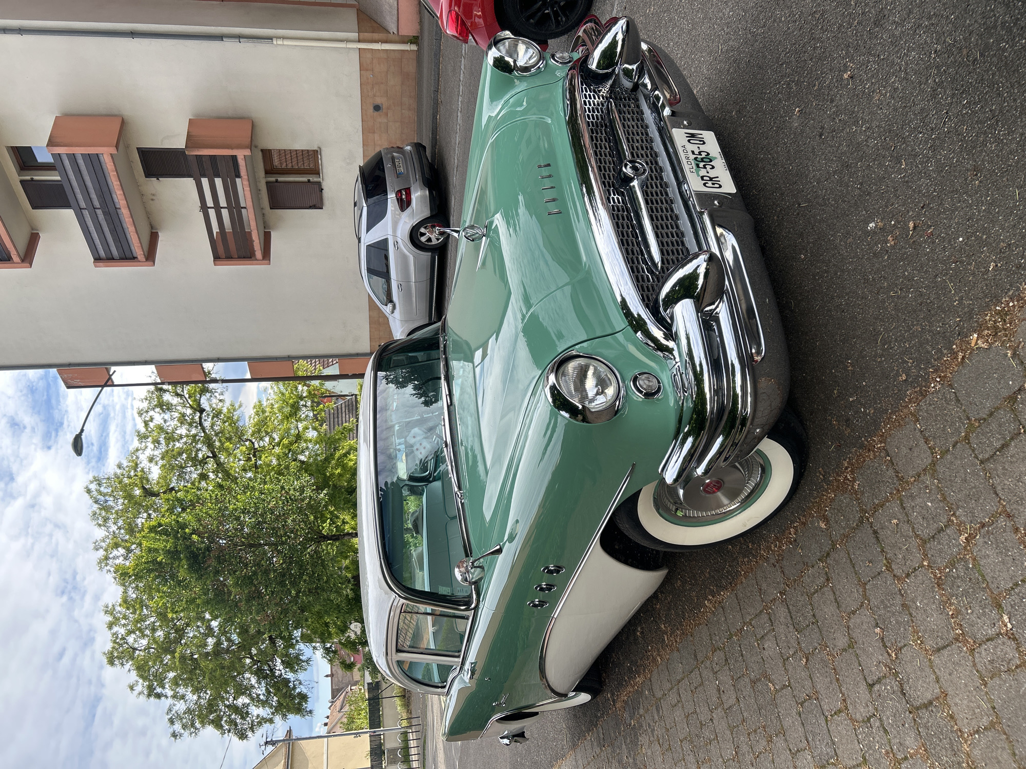 The image shows a vintage two-tone green and white Buick parked on a brick-paved street. The car has a Florida license plate that reads "GR-565-QM." In the background, there are buildings with distinctive window shutters, a silver SUV, and a red car. A large tree is visible to the left. The city is Strasbourg, France.