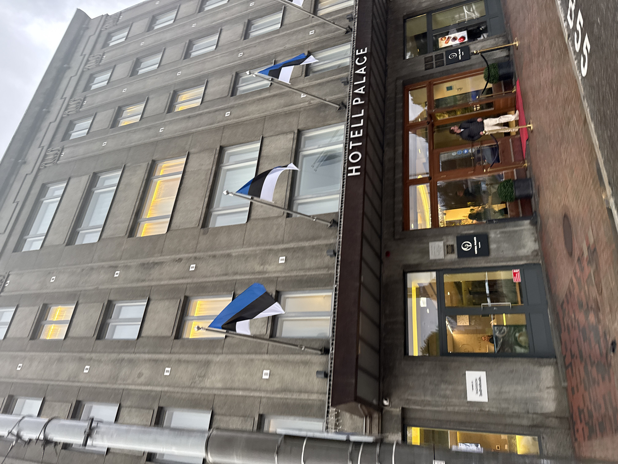 The image shows the exterior of the Hotel Palace in Tallinn, Estonia. The hotel is a tall, gray building with numerous windows, some of which are illuminated with yellow light. Three Estonian flags are prominently displayed on flagpoles attached to the building's facade. The hotel's name, "HOTELL PALACE," is visible above the entrance. The entrance area features glass doors, a red carpet, and stanchions. A man in a dark jacket and light pants is standing on the red carpet outside the entrance. The street in front of the hotel is paved with red brick and asphalt. The sky is overcast, suggesting a cloudy day.